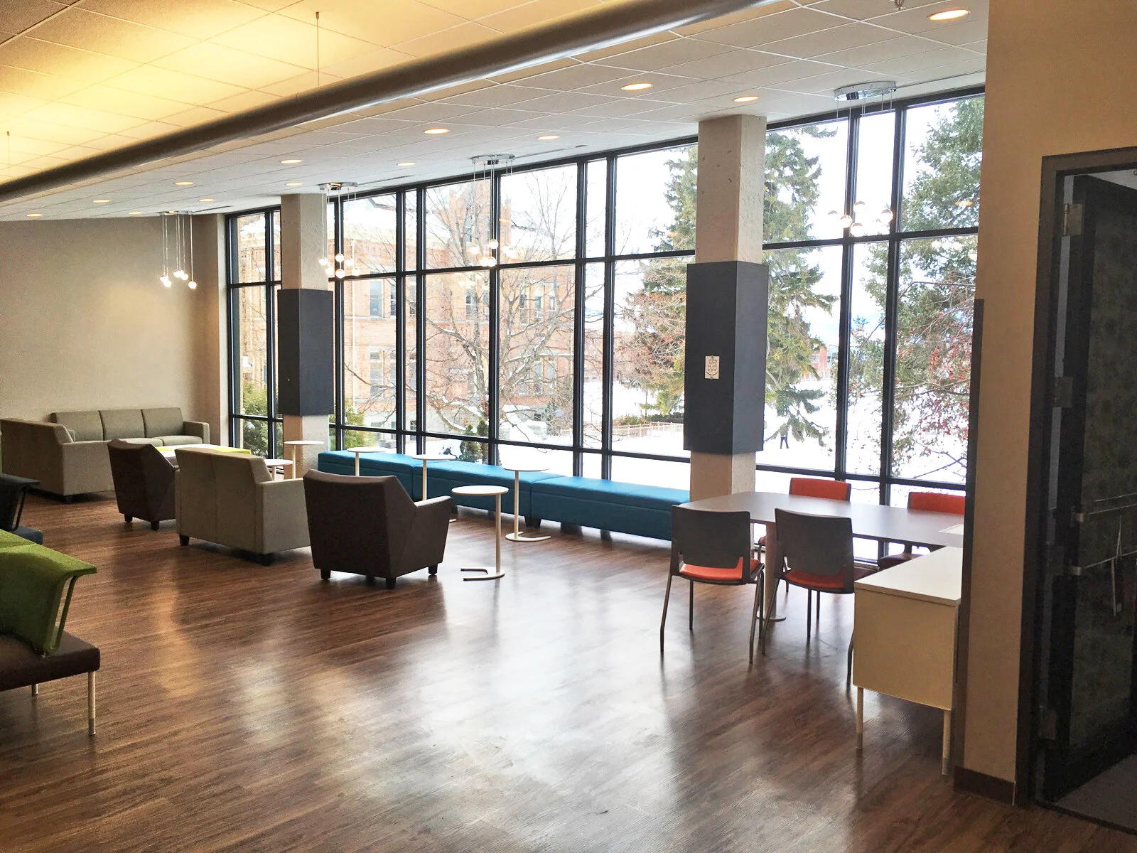 A spacious room with large floor-to-ceiling windows showing outside trees and buildings. There are colorful chairs, sofas, and tables arranged across the room, with a modern design and wooden flooring. The room is well-lit, with pendant lights hangin