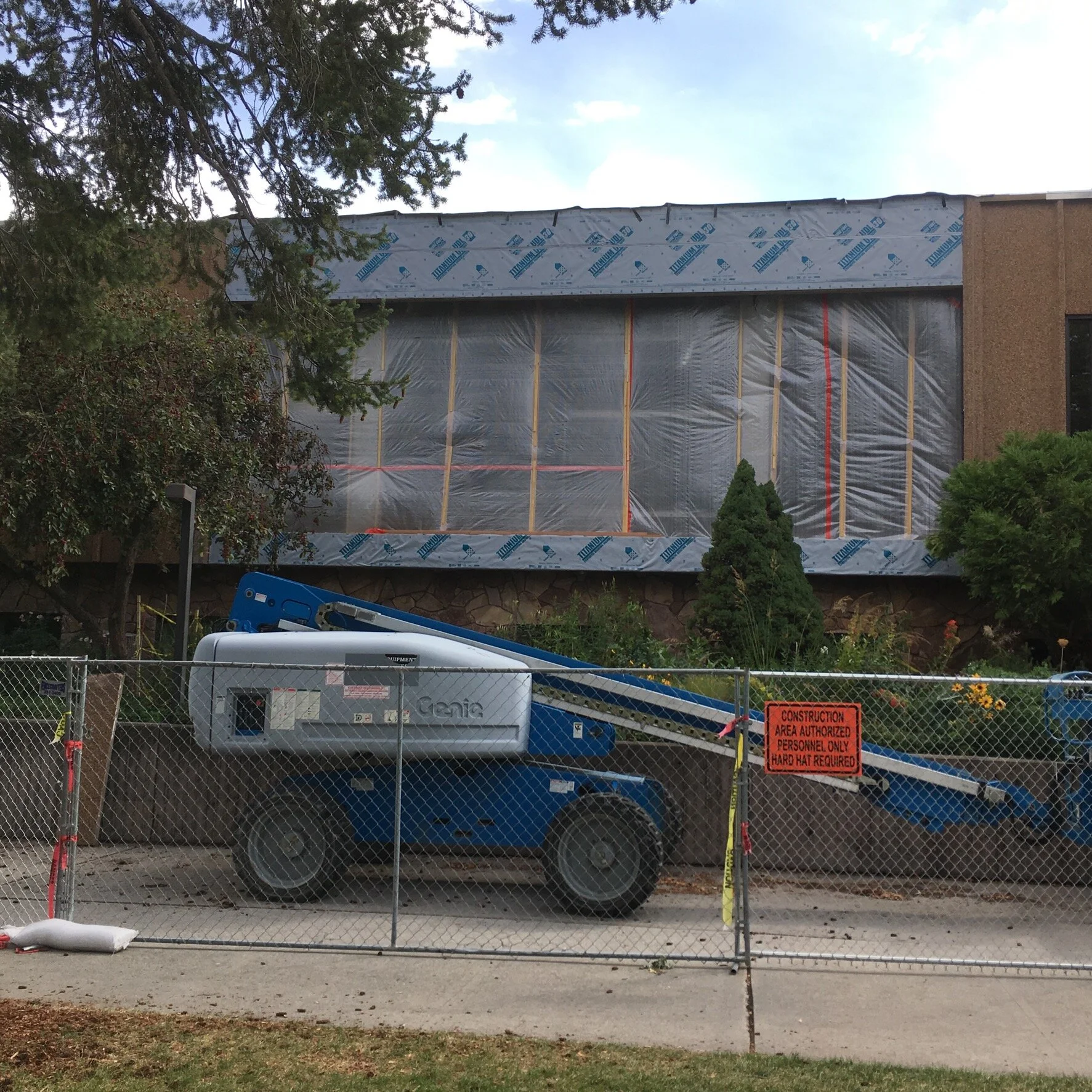 Construction site in front of a building with plastic-covered windows, a blue and white lift parked behind a fence with a sign that says 'Construction area authorized personnel only, hard hat required'.