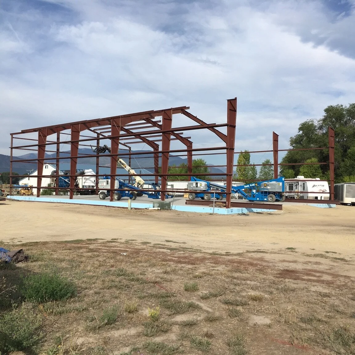 Steel framework of a building under construction with construction vehicles and trailers in the background, and mountains and trees in the distance.