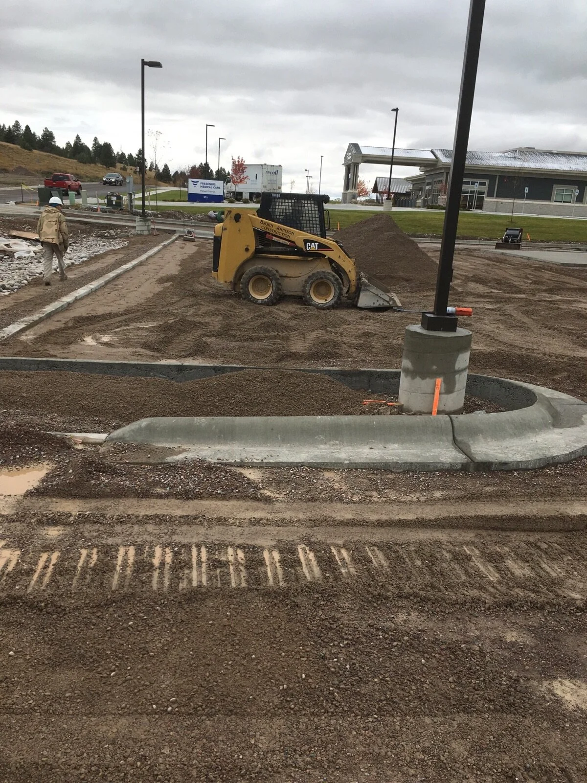 Construction site with a small yellow skid-steer loader moving dirt, a worker in a helmet walking nearby, and unfinished pavement and curbs under a cloudy sky.