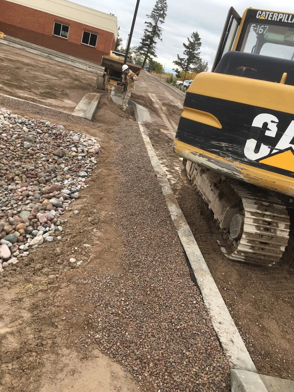 Construction site with worker leveling gravel and compacting the ground near a sidewalk, with a yellow excavator on one side and a building in the background.