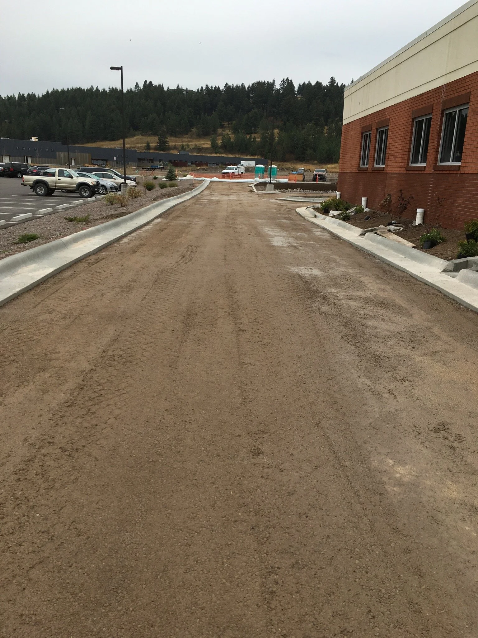 A dirt driveway or parking area near a brick building under construction, with soil and curb edges on either side, and a parking lot with cars in the background.