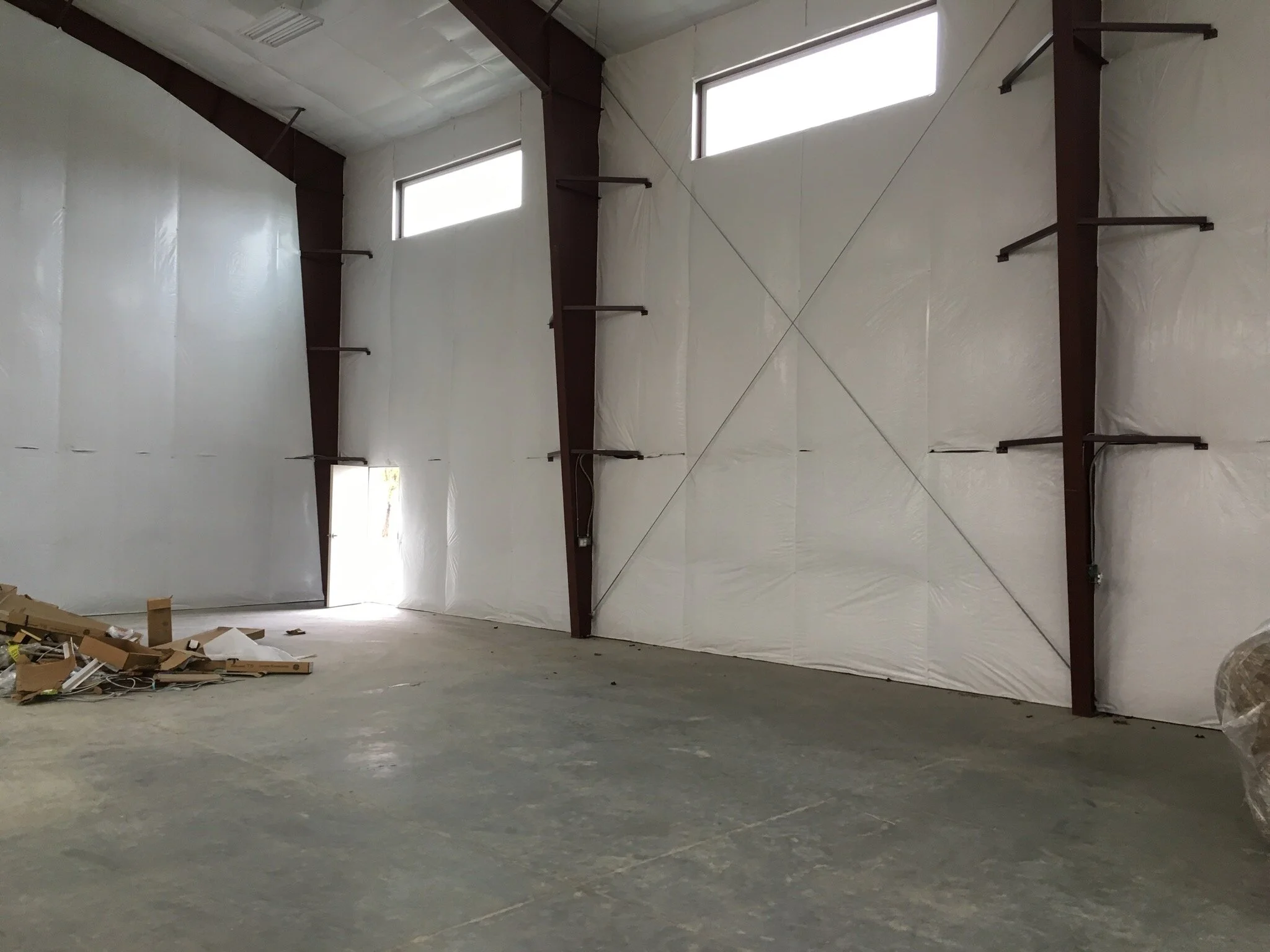 Empty warehouse with white insulation on walls, high windows, and metal support beams, with some construction materials on the floor.
