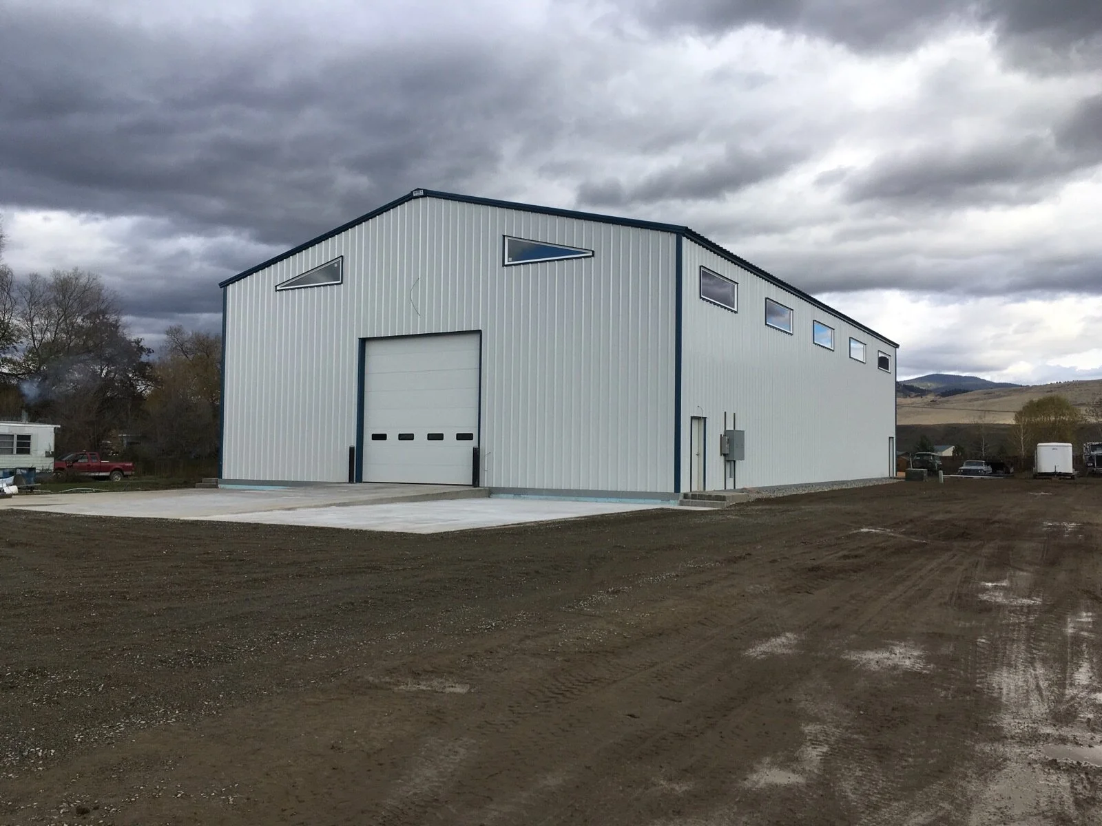 Large light gray metal building with a white garage door and small triangular top windows, set in a rural landscape under cloudy sky.