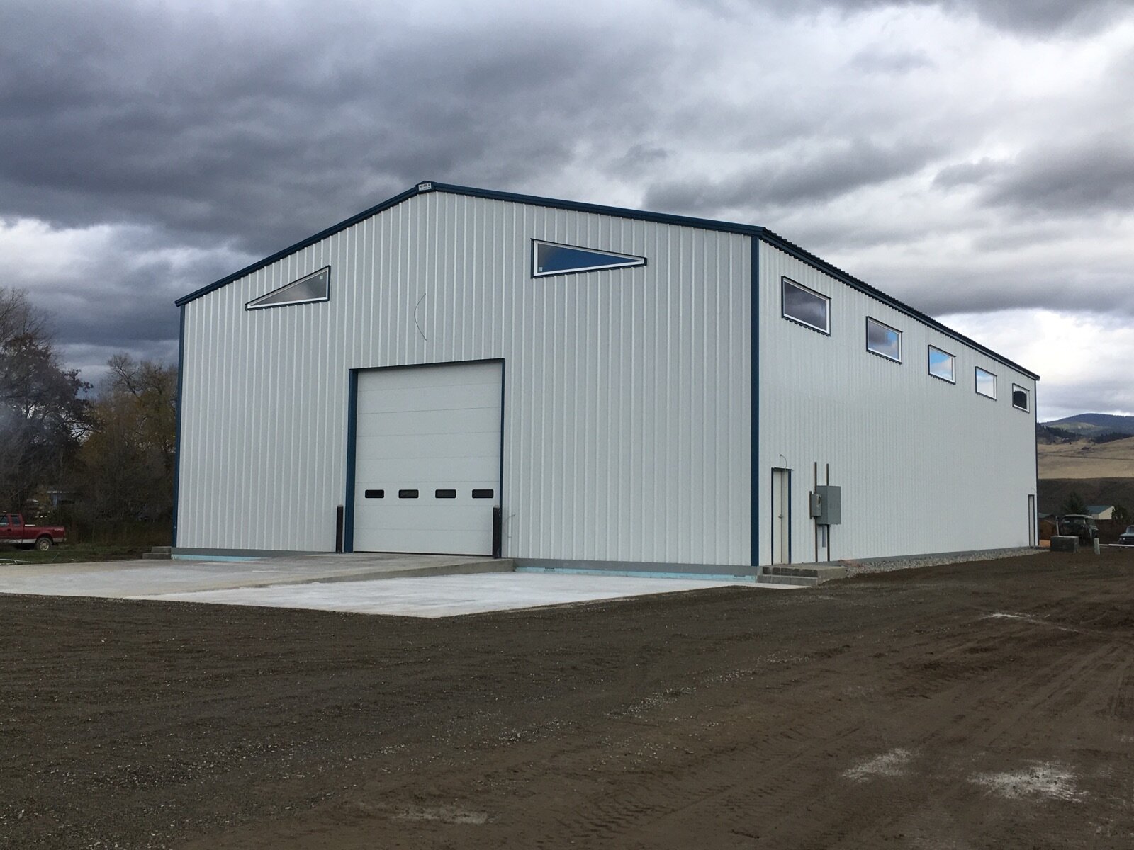 A large, white metal building with a garage door and small, triangular windows, situated on a dirt lot with overcast skies overhead.