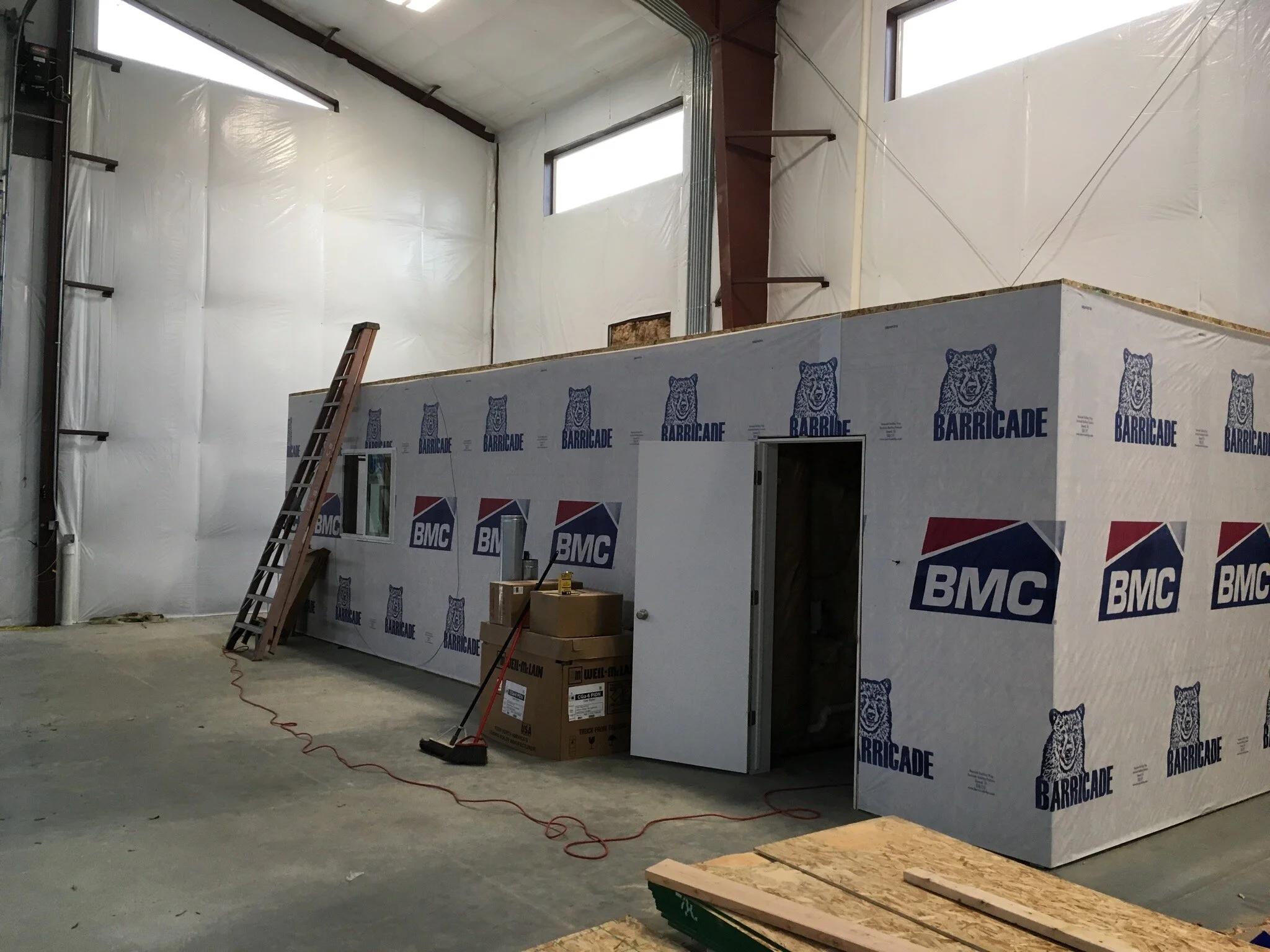 Interior of a building under construction with a partially finished wall covered in insulation material. There is a ladder leaning against the wall, and construction tools and boxes are scattered on the floor.