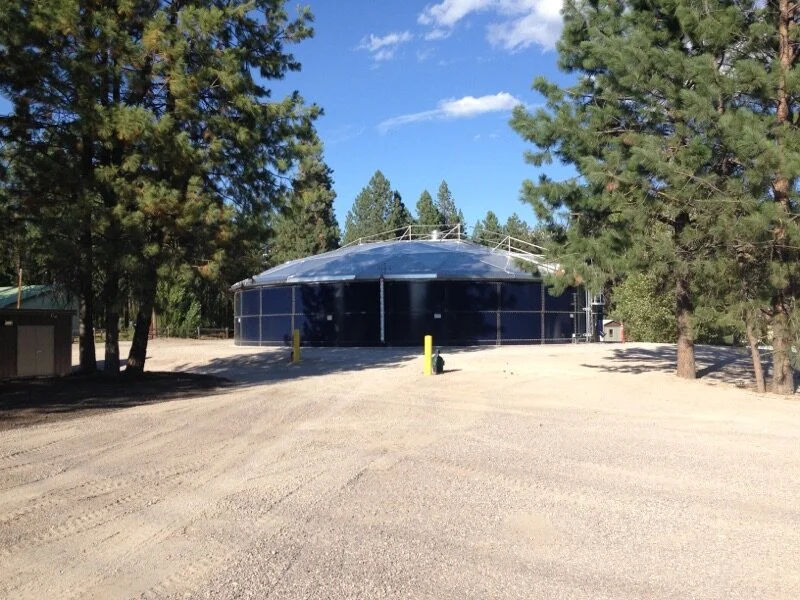 A large cylindrical water storage tank with a blue exterior surrounded by trees and some utility poles in a rural area.