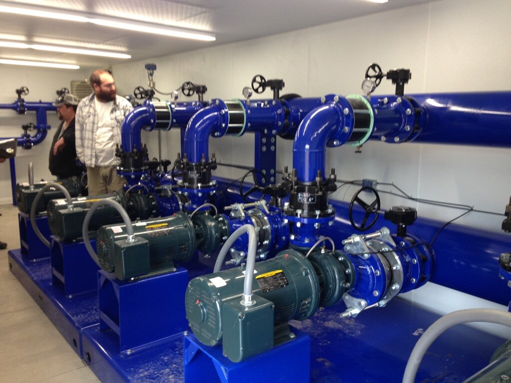 Two men examining a large mechanical room with blue pumps and pipes.