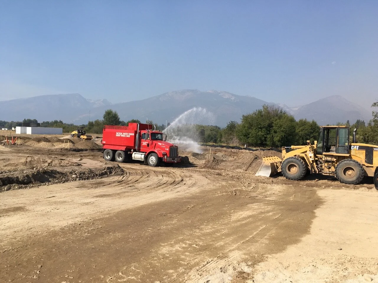 Construction site with a red water tanker truck spraying water and a yellow bulldozer moving dirt, with mountains in the background.