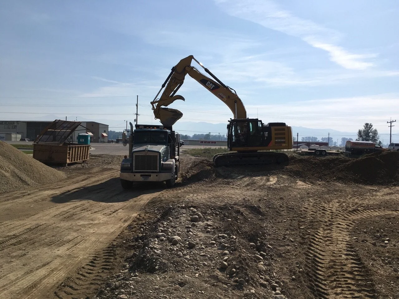 An excavator loading dirt into a dump truck at a construction site under a blue sky.