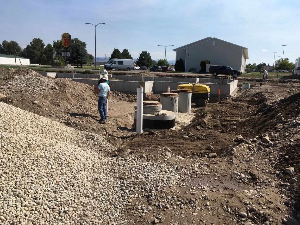 Construction site with workers, dirt piles, and underground utility components with buildings and vehicles in the background.