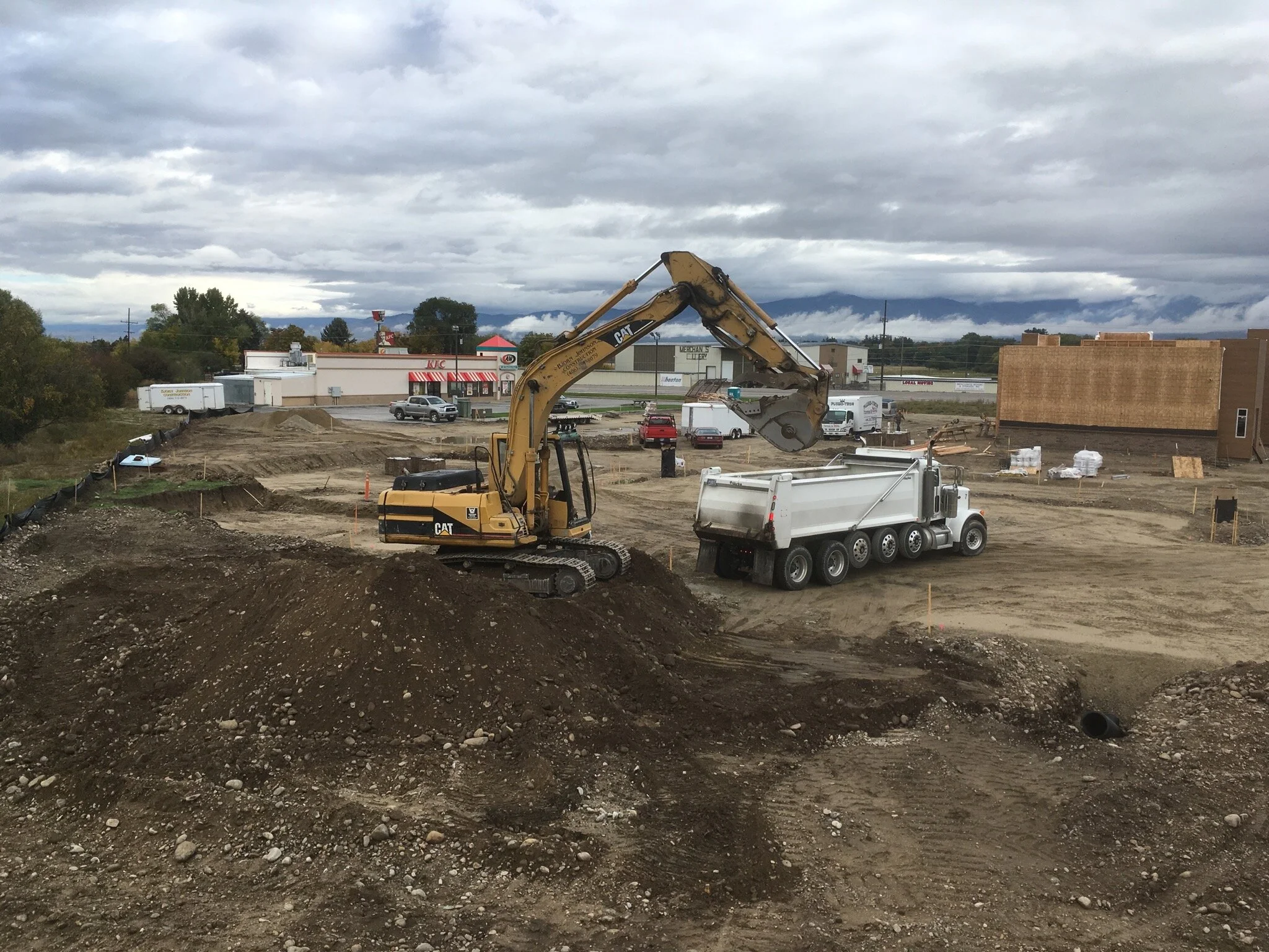 Construction site with a yellow excavator loading dirt into a white dump truck, with buildings and cloudy sky in the background.