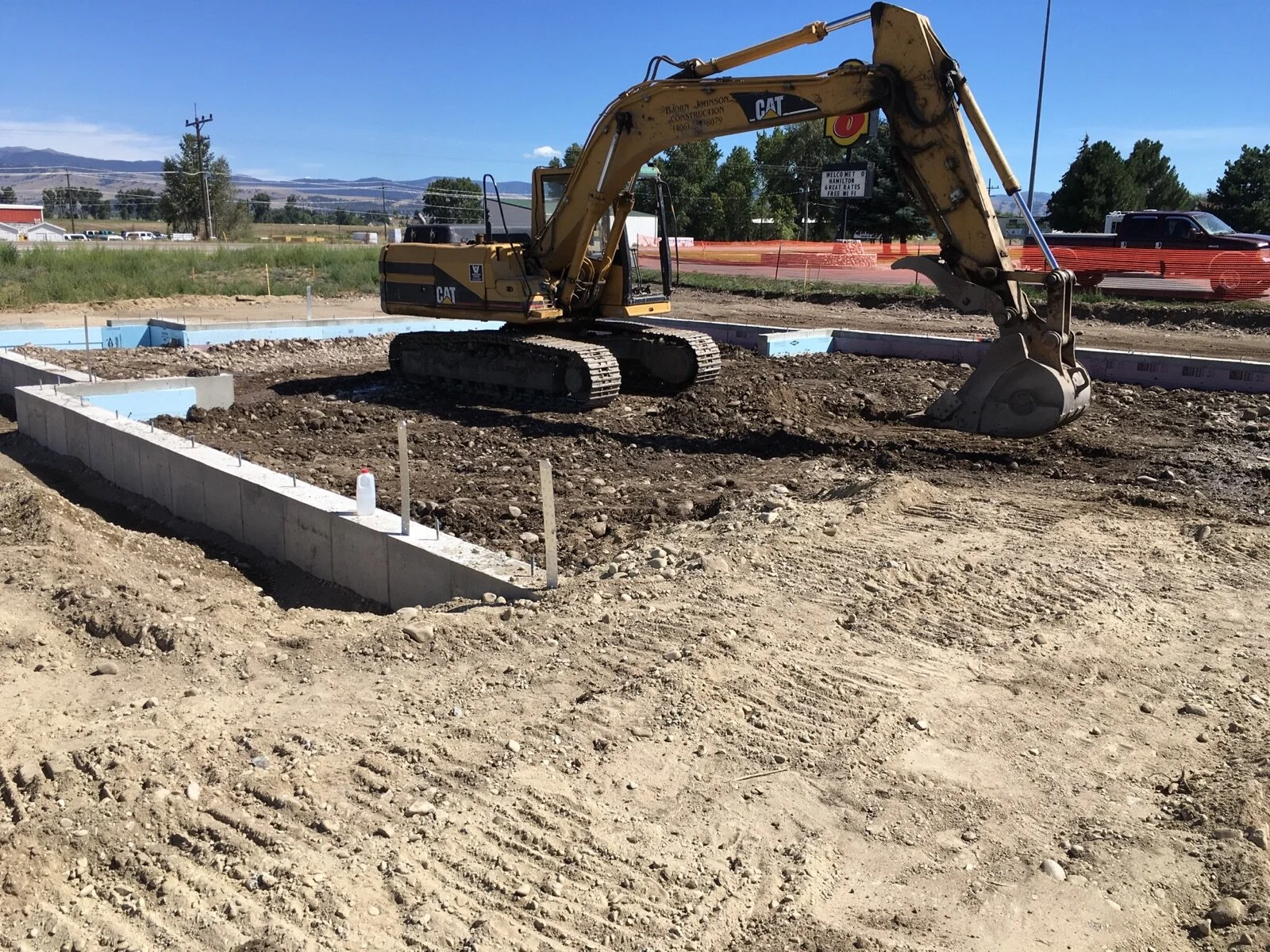 Construction site with a yellow Caterpillar excavator digging in dirt, concrete foundation walls, and blue insulation panels on the ground, under a clear blue sky.