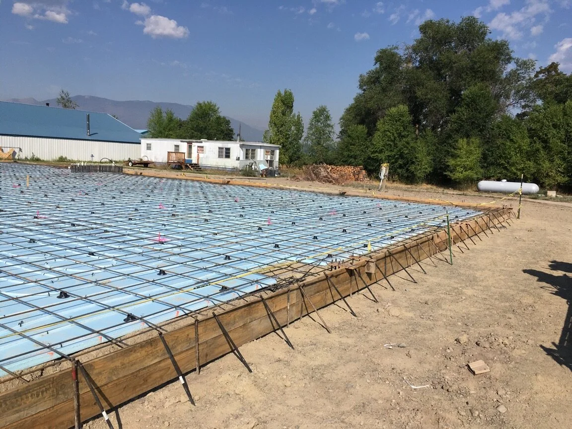 Construction site with a grid of metal rebar and plastic spacers prepared for pouring concrete, surrounded by dirt, with trees and a building in the background under a blue sky.