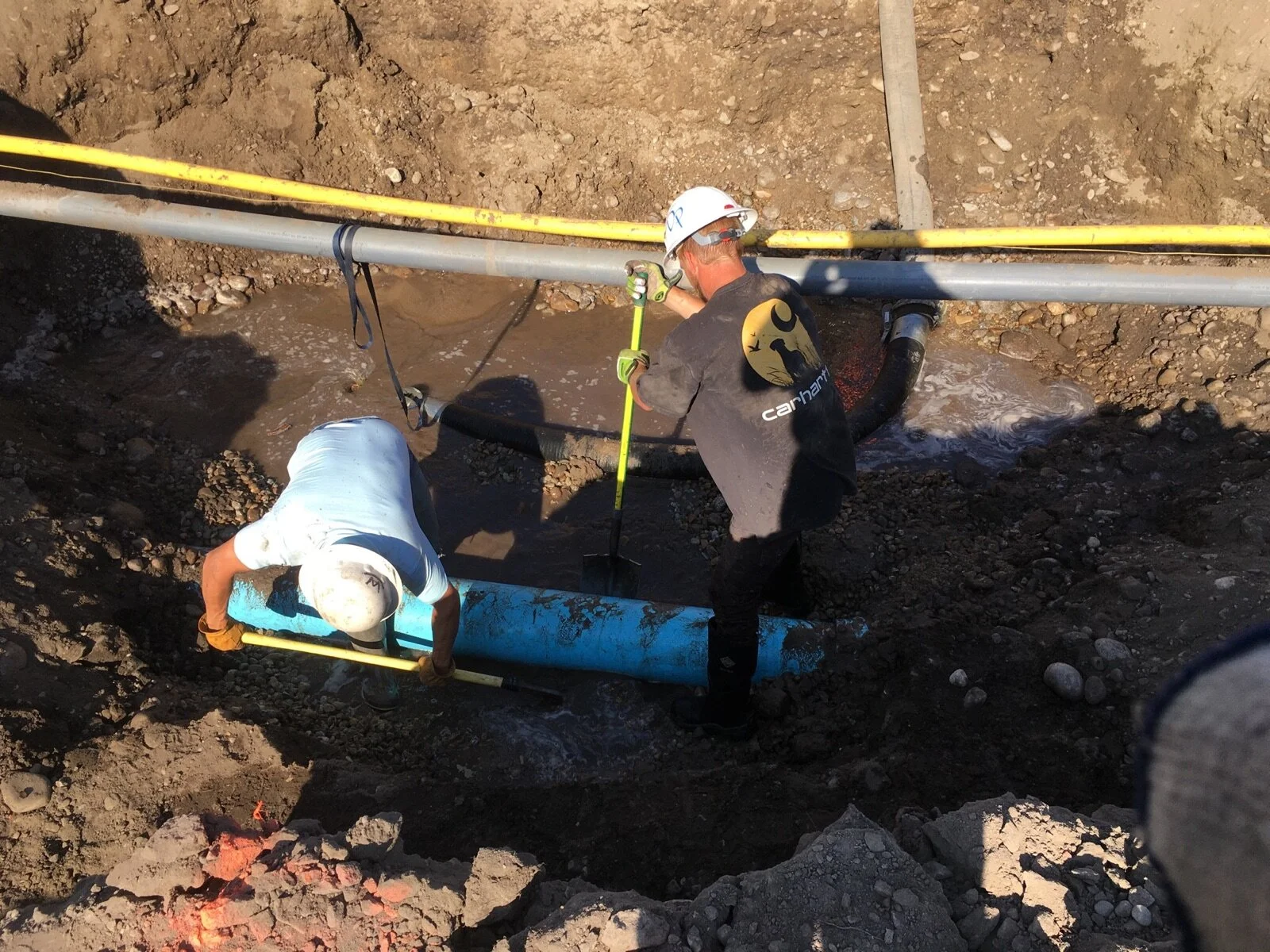 Two workers are installing or repairing underground pipes in a trench, one wearing a white shirt and helmet, the other in a dark shirt with a helmet, using tools to connect the pipes.