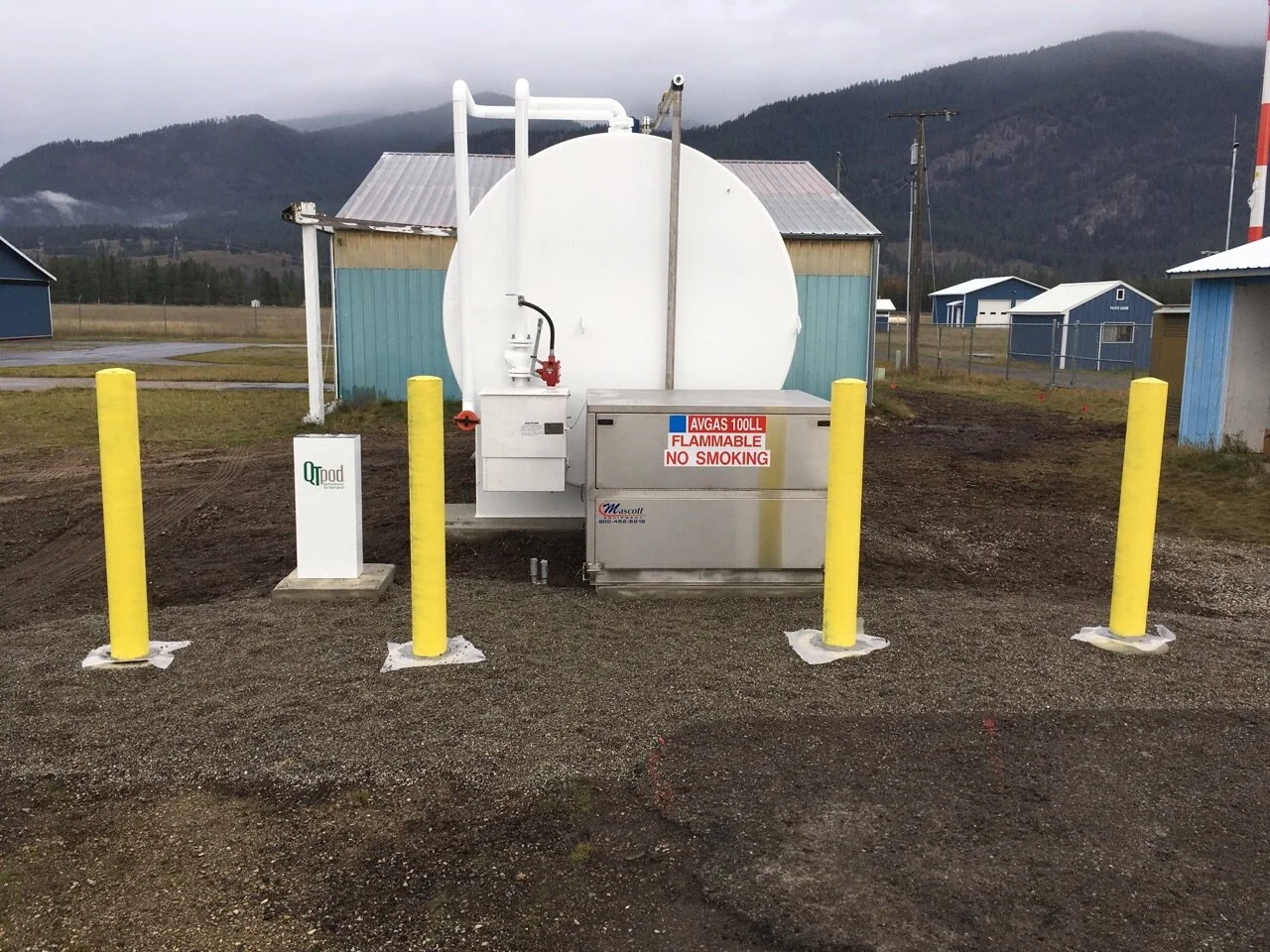 Propane tank station with yellow safety bollards and a warning sign, in a rural area with small buildings and mountains in the background.