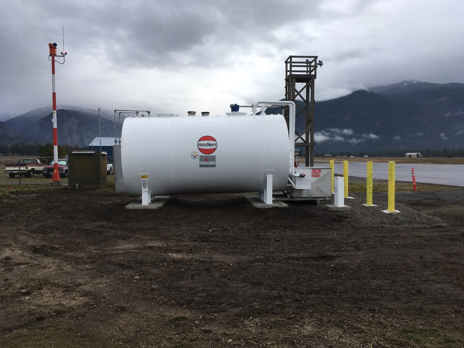 White tank labeled 'modern' with warning signs, positioned outdoors on dirt, with yellow safety posts nearby, mountain range and cloudy sky in the background.