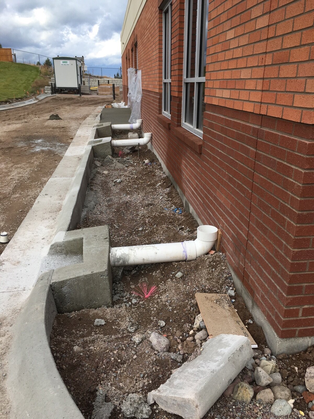 Construction site showing a brick building with window, outdoor drainage pipes along the foundation, and dirt yard with construction materials and equipment.