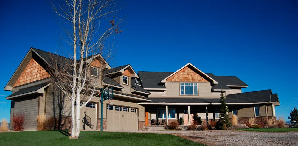 Large modern house with multiple gabled roofs, gray siding, brick accents, and a two-car garage, set against a clear blue sky, with a tall leafless tree and landscaped yard.