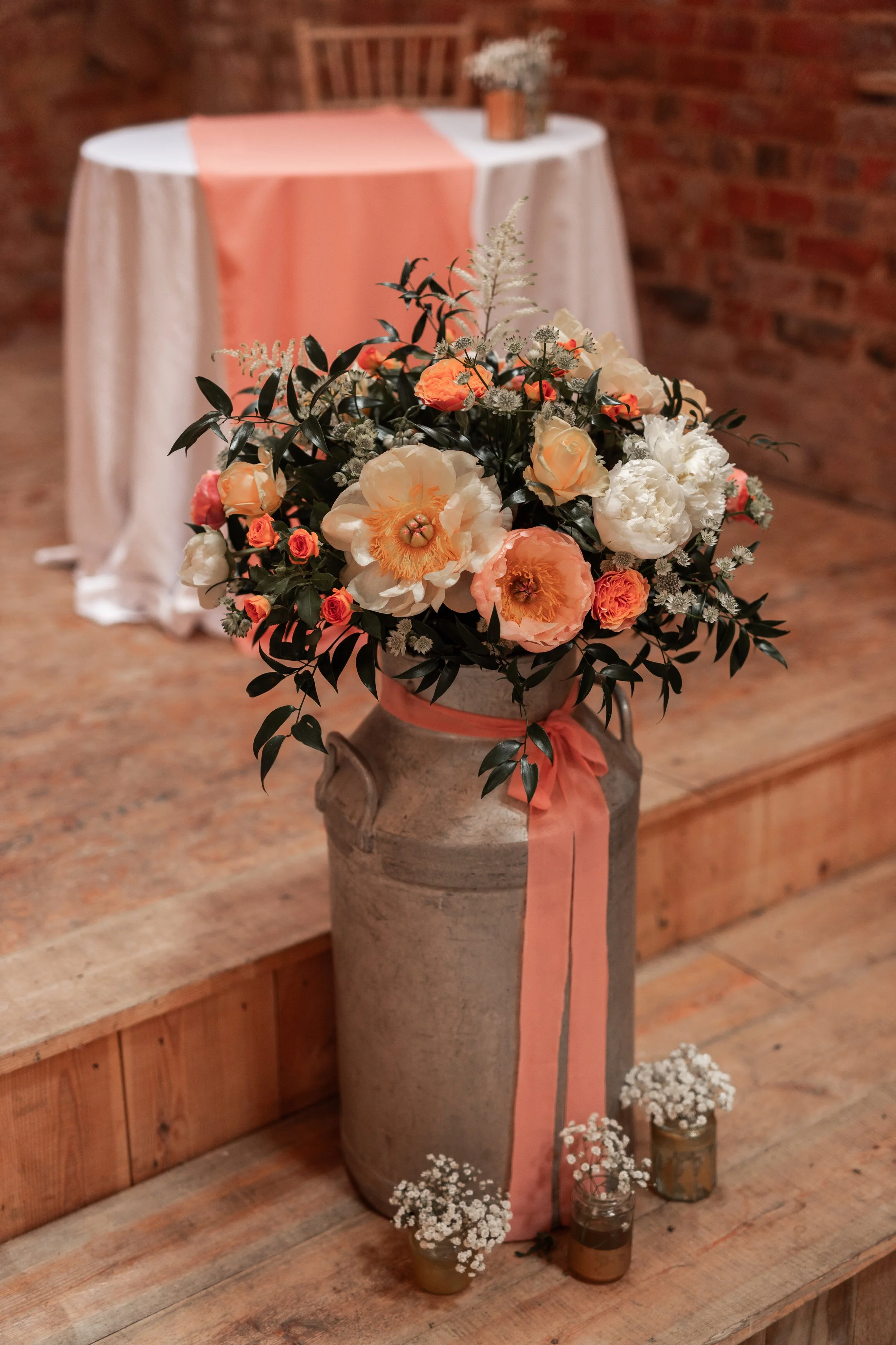 A large floral arrangement with white, peach, and pink flowers in a tall, rustic vase decorated with a pink ribbon, placed on a wooden surface.