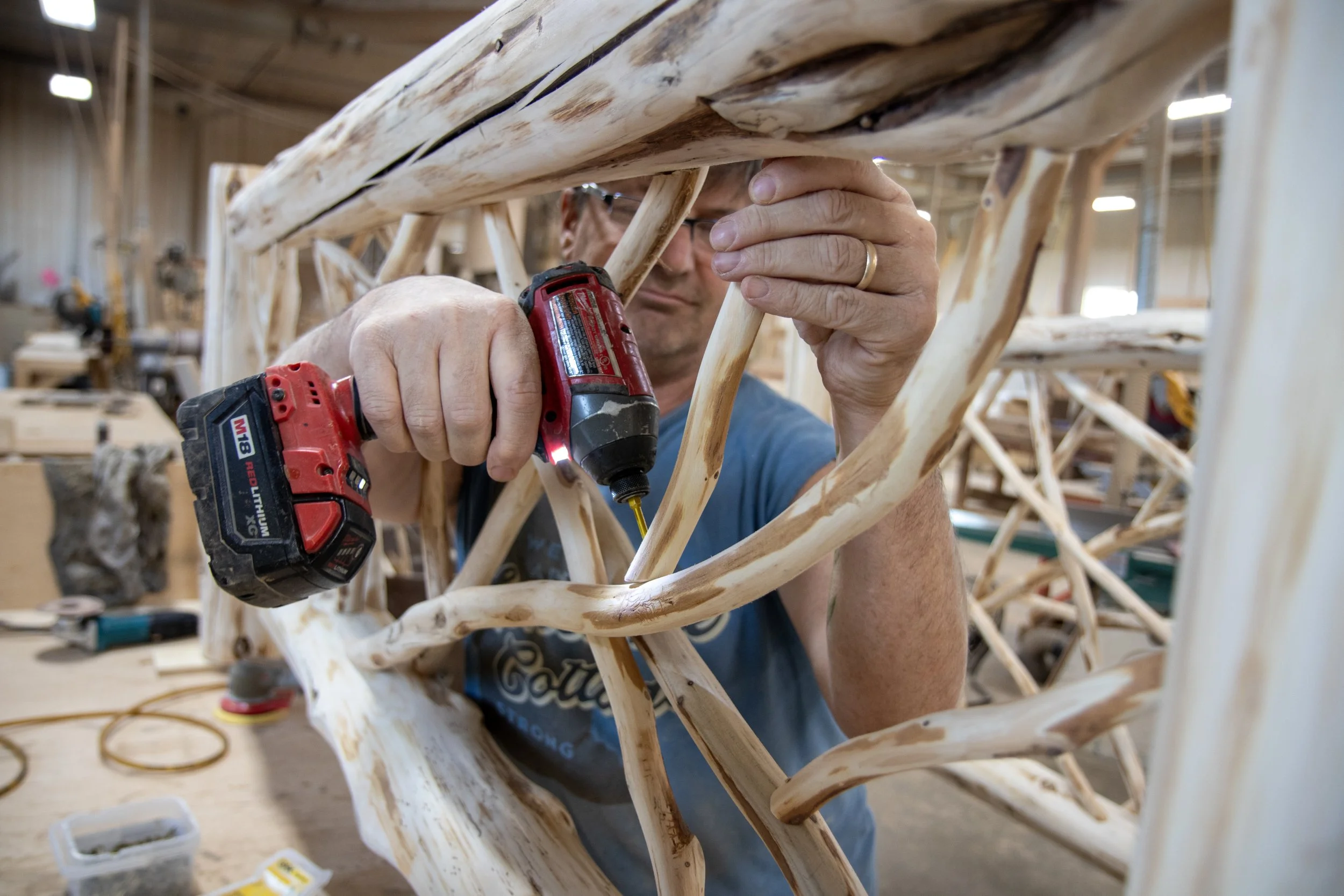 A man working on a wooden chair frame in a woodworking shop, using a power drill.