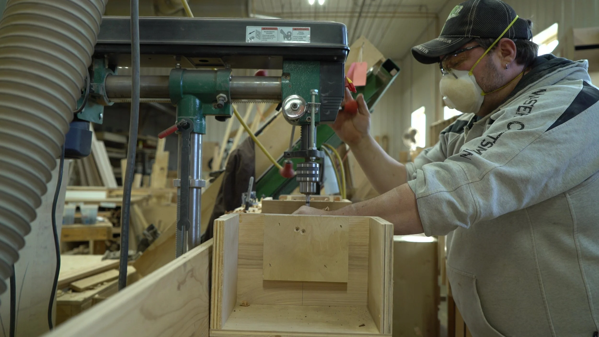 A person wearing safety glasses, a face mask, and a cap is using a drill press to work on a piece of wood in a woodworking shop, with wooden frames and tools visible in the background.