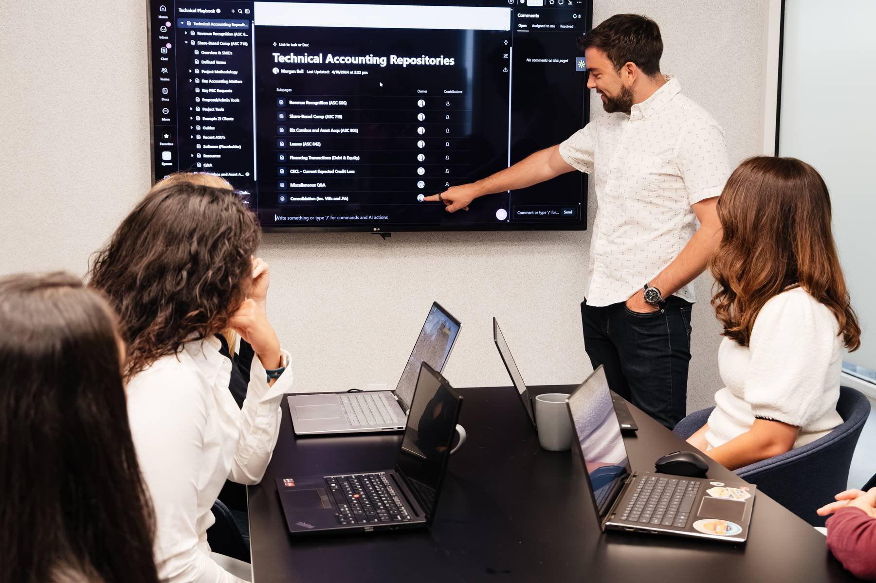 A man is giving a presentation on a large screen that displays a digital interface titled 'Technical Accounting Repositories.' Four women are seated at a black conference table with laptops open, listening to the presentation.