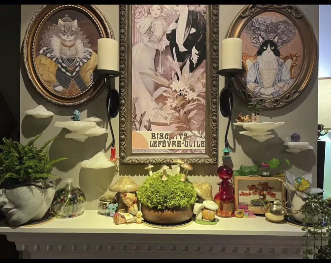 Decorative display with framed artwork of cats dressed in vintage clothing, surrounded by wall-mounted mushroom shelves with small objects, candles, and plants, all on a white mantelpiece.