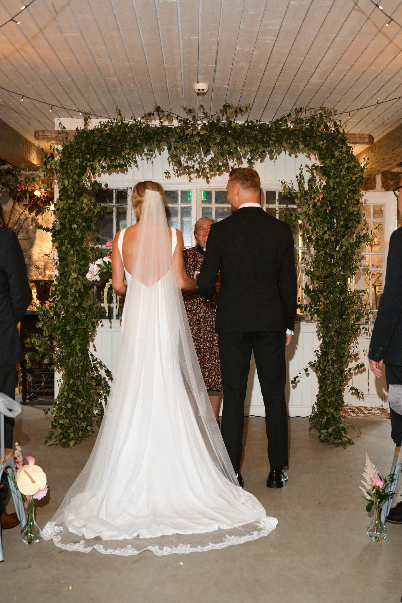 En brud och en brudgum står under ett blomsterdekorerat bröllopsark av grön bladverk, och håller varandras händer under ett bröllopsceremoni.