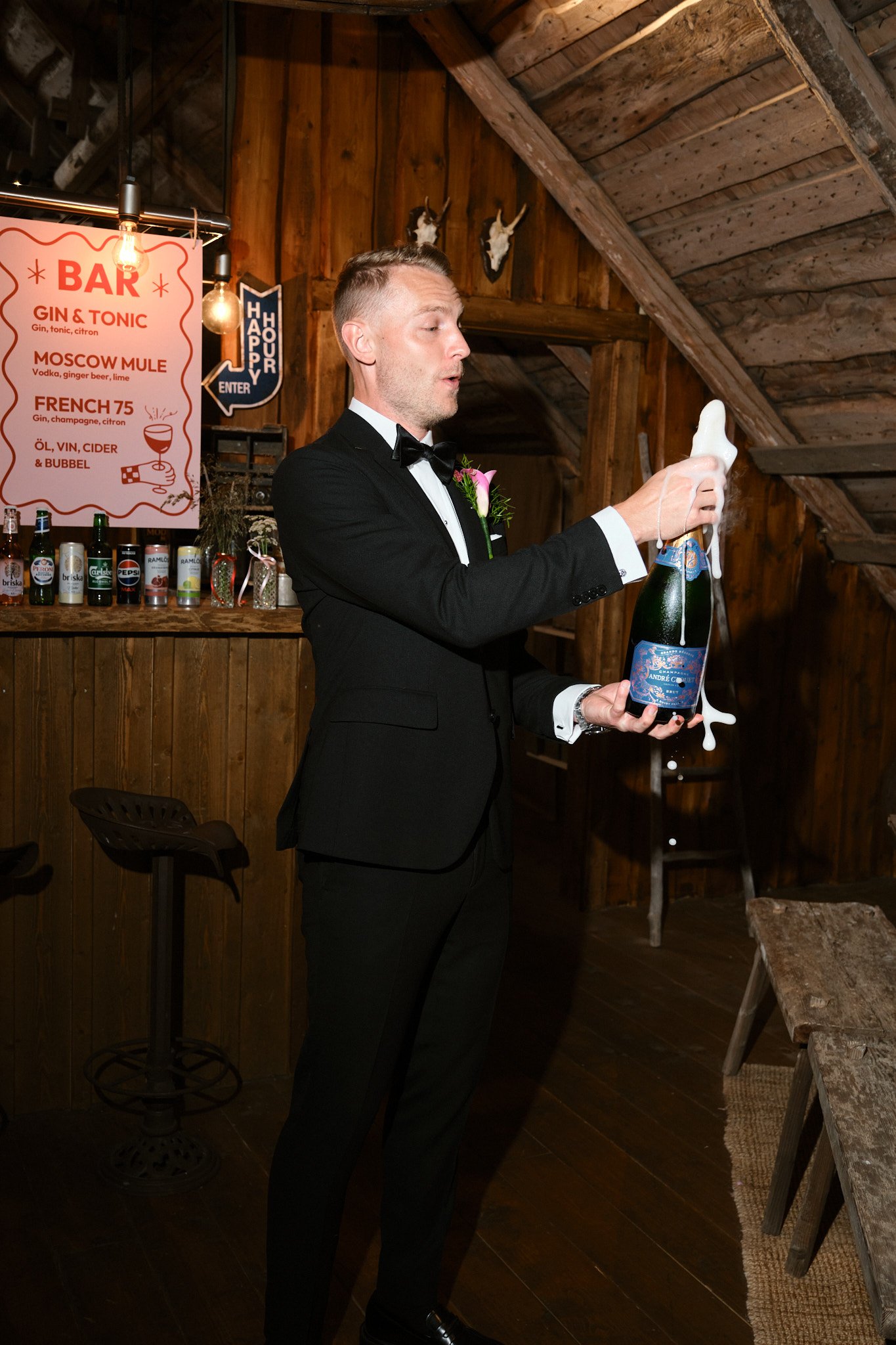 En man i svart tuxedo med svart fluga öppnar en champagneflaska med skum som flyter ner.