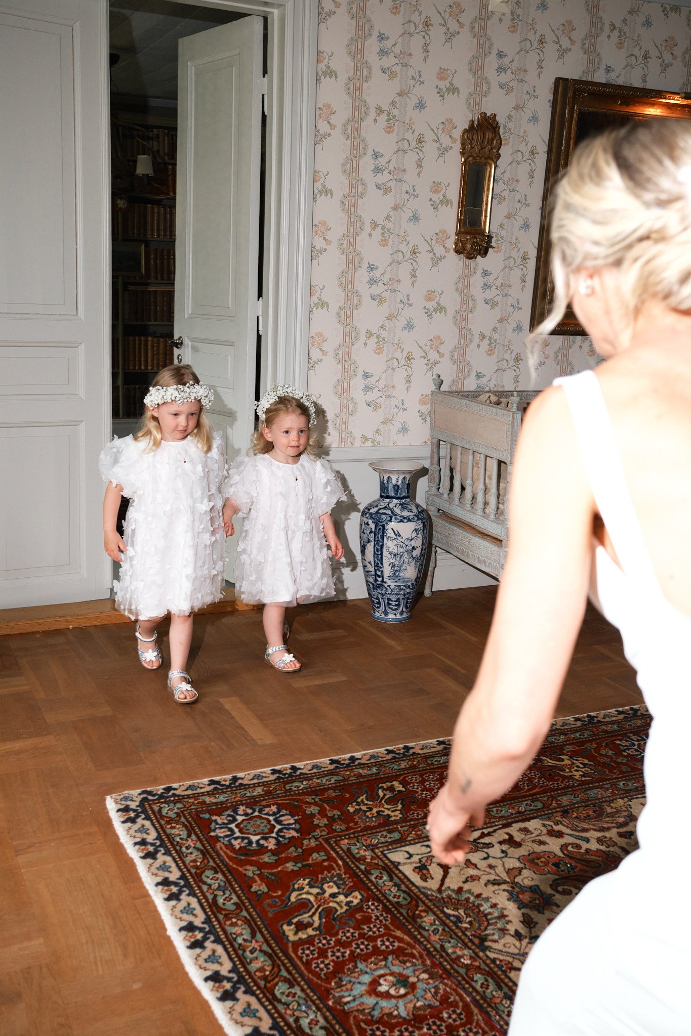 Två små flickor i vita klänningar och blomsterkransar på huvudet går in i ett rum, en vuxen kvinna i vit klänning står i närheten, inomhus med blommig tapet och ett kinesiskt blåvitt vaser.