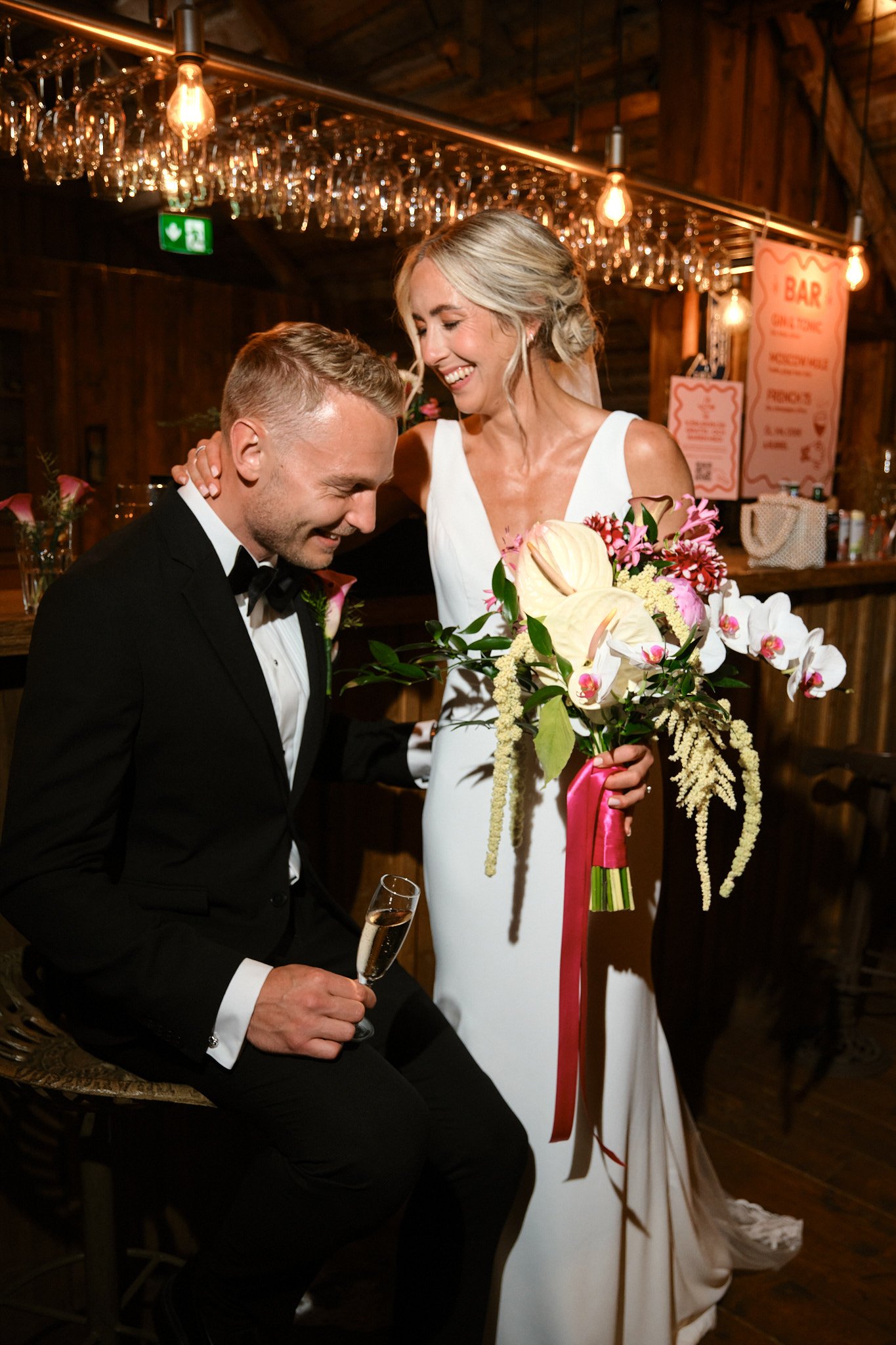 En brud och en groom som ler och skrattar, bruden håller en stor blombukett, de är i en bar eller restaurang med varmt belysning och träväggar.