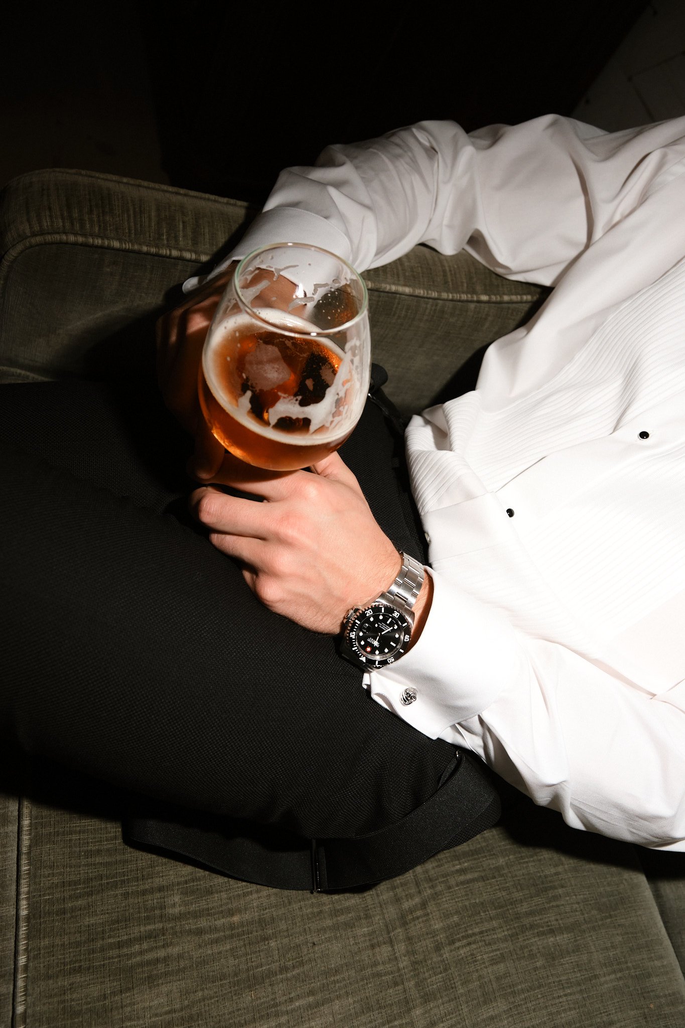 Person som bär vit skjorta och svart byxor, sitter i en soffa, håller i ett glas öl.