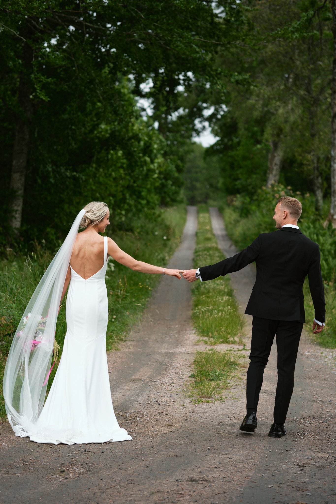 Brudpar på en skogsstig, hon i vit brudklänning och han i svart kostym, håller varandra i handen.