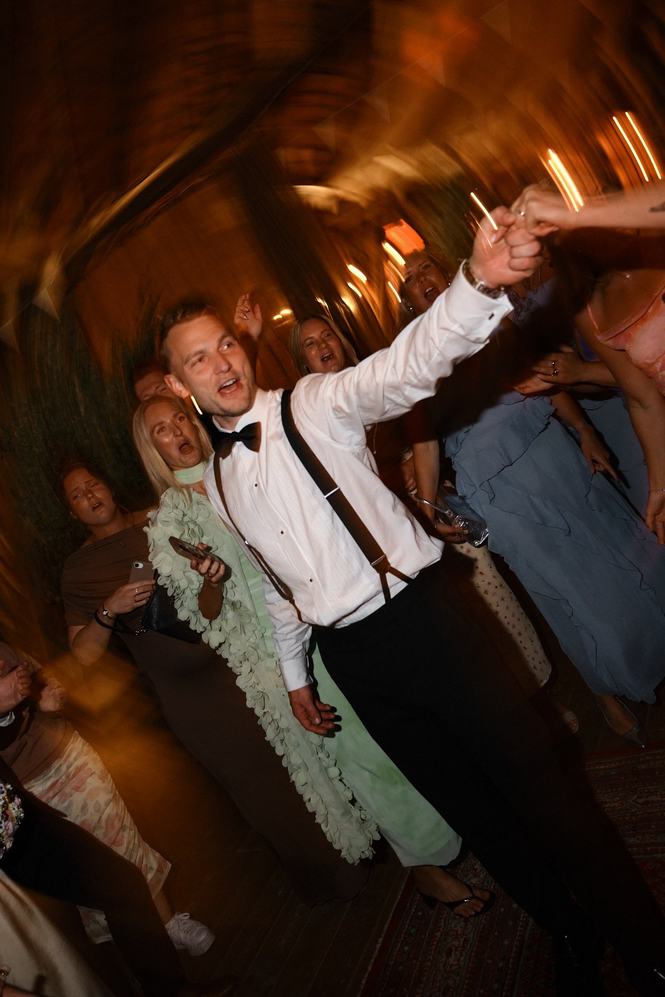 En grupp människor dansar och har kul på en fest, med en man i vit skjorta och svart fluga som leder dansen.