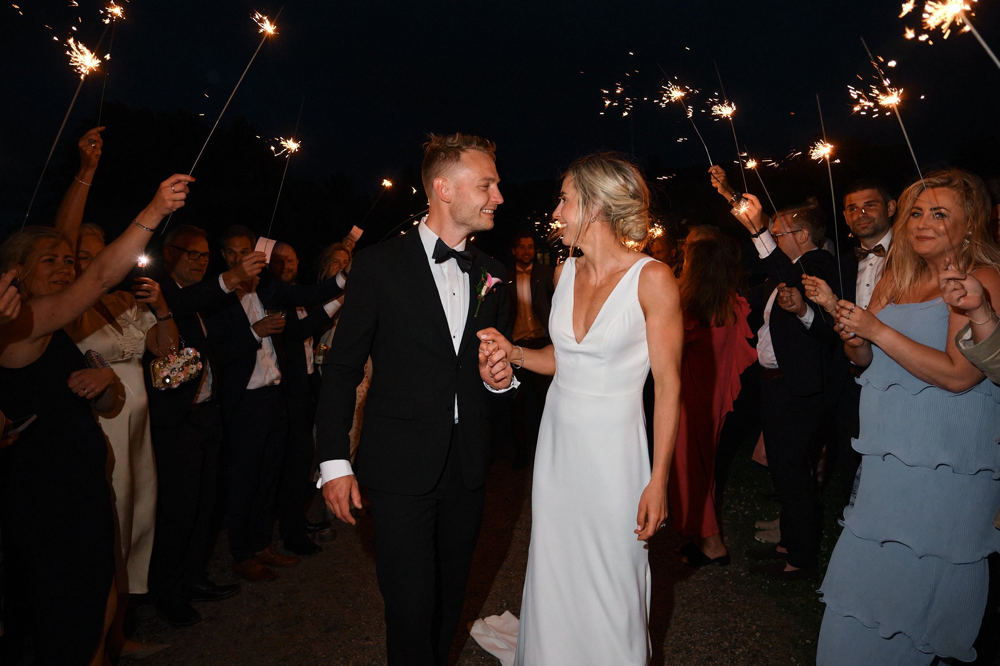 Brudpar dansar under fyrverkerier omgiven av vänner och familj vid nattbröllop.