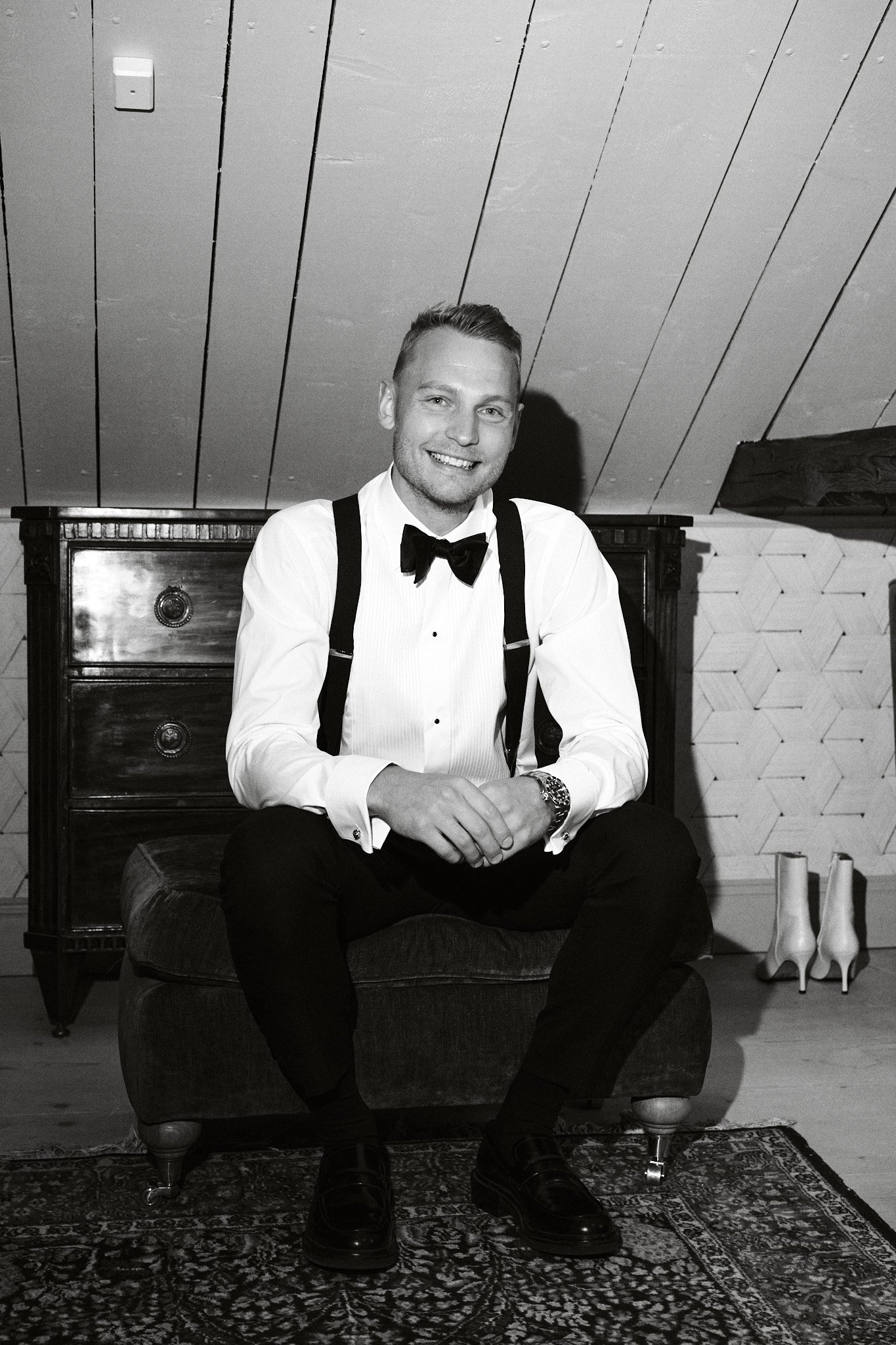 En glad ung man i formell klädsel inklusive vit skjorta, svart fluga och svarta byxor, sitter på en fåtölj i ett rum med tak av träpanel. Bredvid honom står ett par vita stövlar.