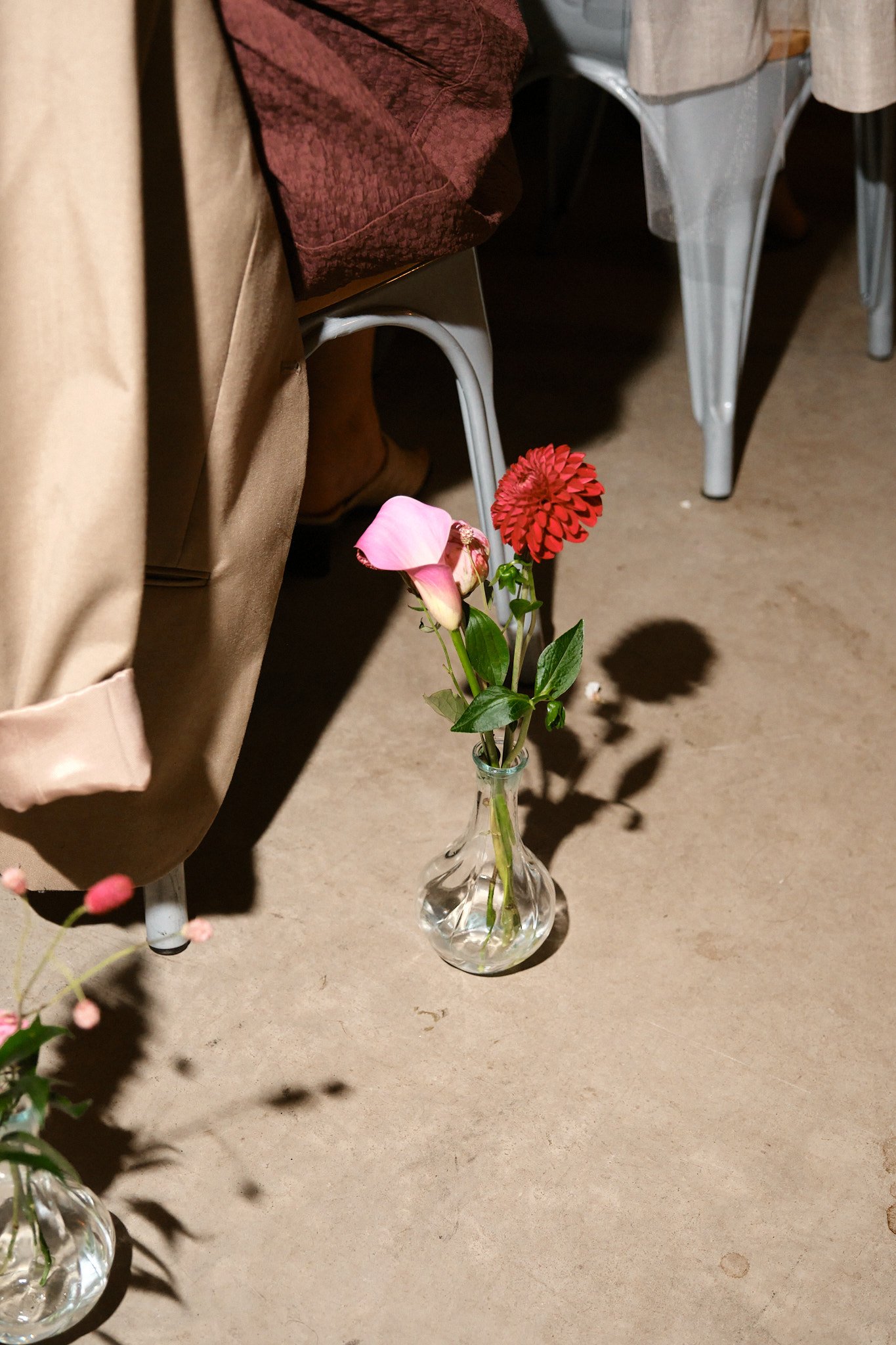 En liten glasvas med tre blommor: en rosa klocka, en mörkröd dahlia och en ljusrosa calla, placerad på ett golv nära en person som sitter på en stol.
