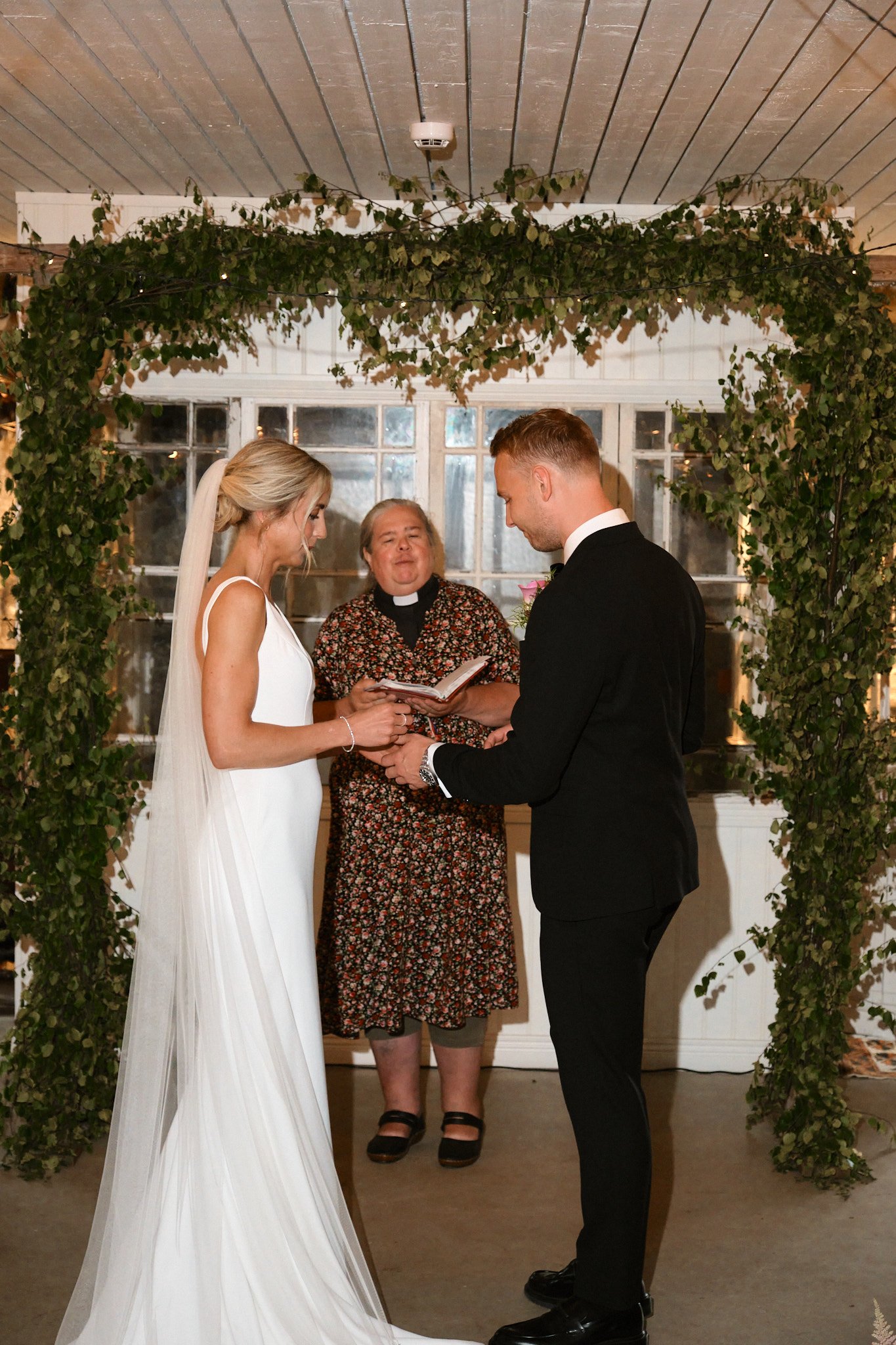 En brud och en brudgum som håller hand under ett bröllopscitatt, med en officiant som läser från en bok, under ett uterum dekorerat med gröna växter.