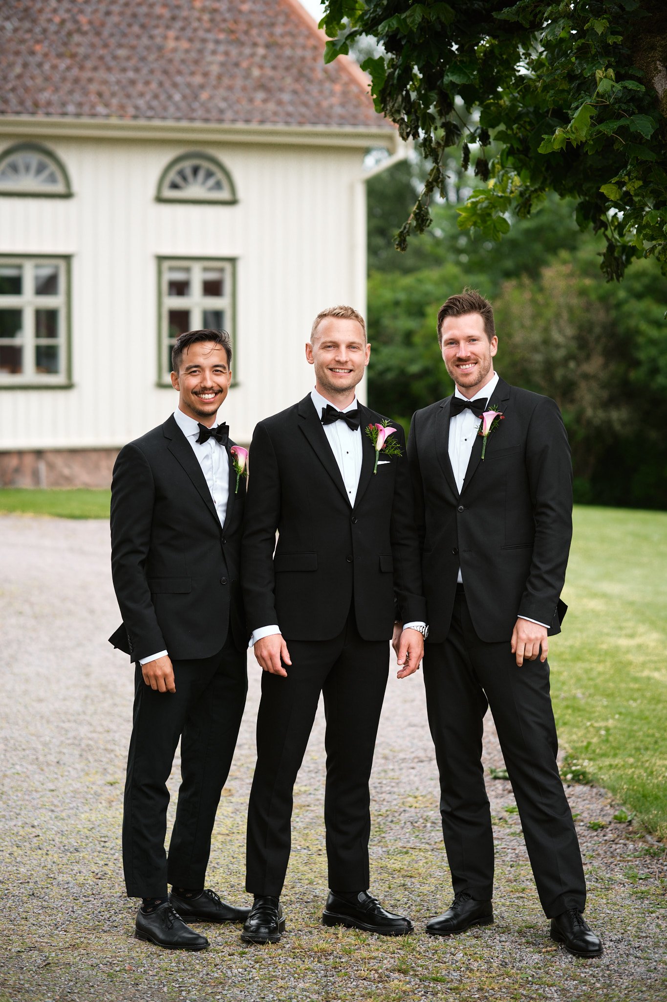 Tre män i svart kostym och fluga, klädda för ett bröllop, står utomhus framför ett vitt hus med gröna fönster och träd.