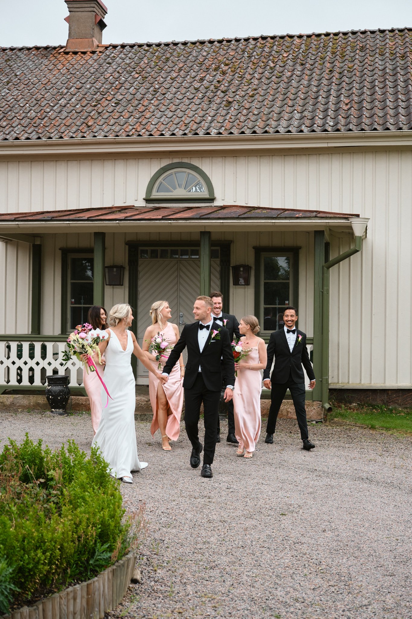 Brudparet med brudtärnor och best man lämnar ett hus efter bröllop. Alla är klädda i formella kläder, och de håller buketter, mitt på en grusuppfart.