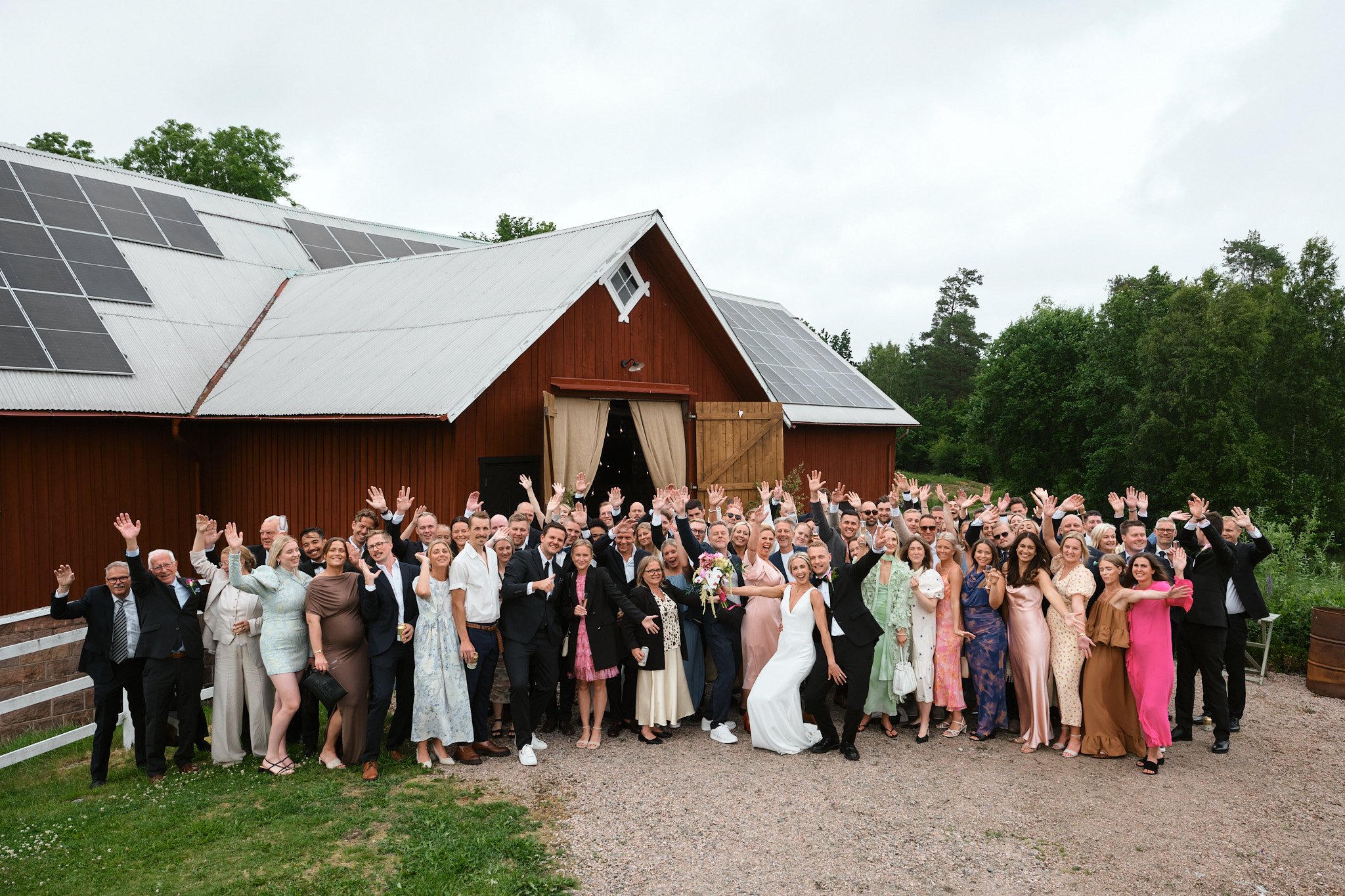 En stor grupp människor på ett bröllop utomhus framför en röd lada, de vinkar och ler mot kameran.