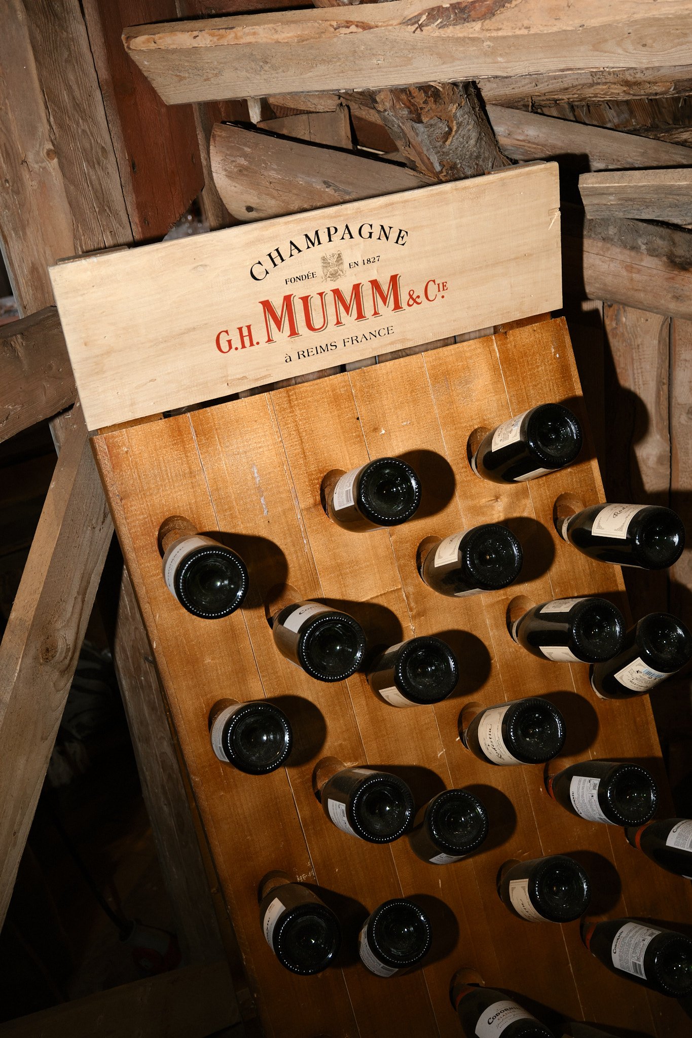En träställning med tomma vinflaskor, märkt med G.H. Mumm & Cie, i en vintappning. Resten av rummet är fullt av trästugor och brädor.