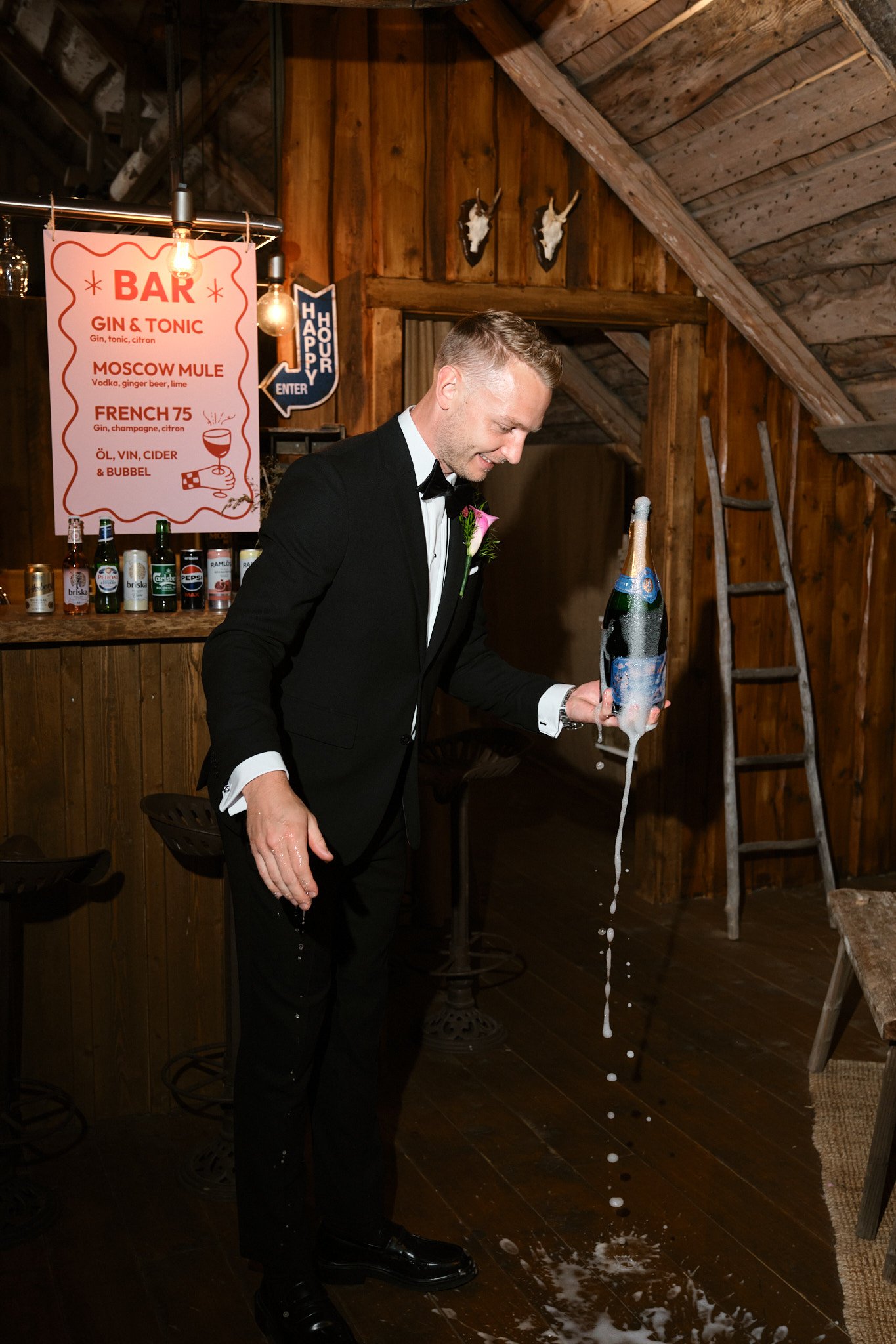 En man i svart tuxedo och vit skjorta som skakar en champagneflaska, vilket orsakar att korken skjuter ut och dryck och foam skvätter. Han står i ett rustikt rum med träväggar och en bar i bakgrunden.
