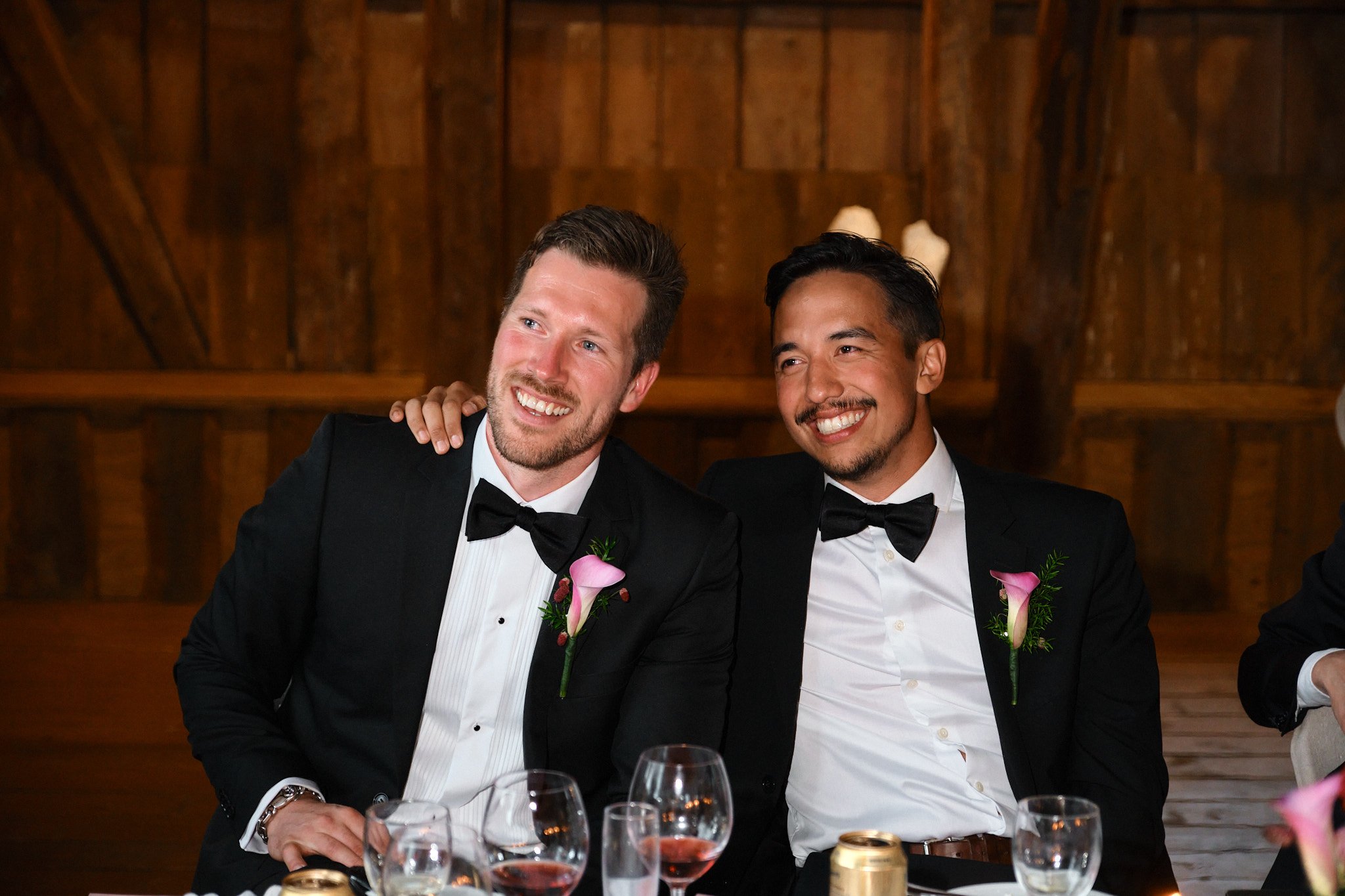 Två män i smoking och korsade knäppta manchetter, med rosa blommor som bröllopsboutonniere, sitter vid ett bord och ler. De ser glada och avslappnade ut, med en trävägg i bakgrunden.
