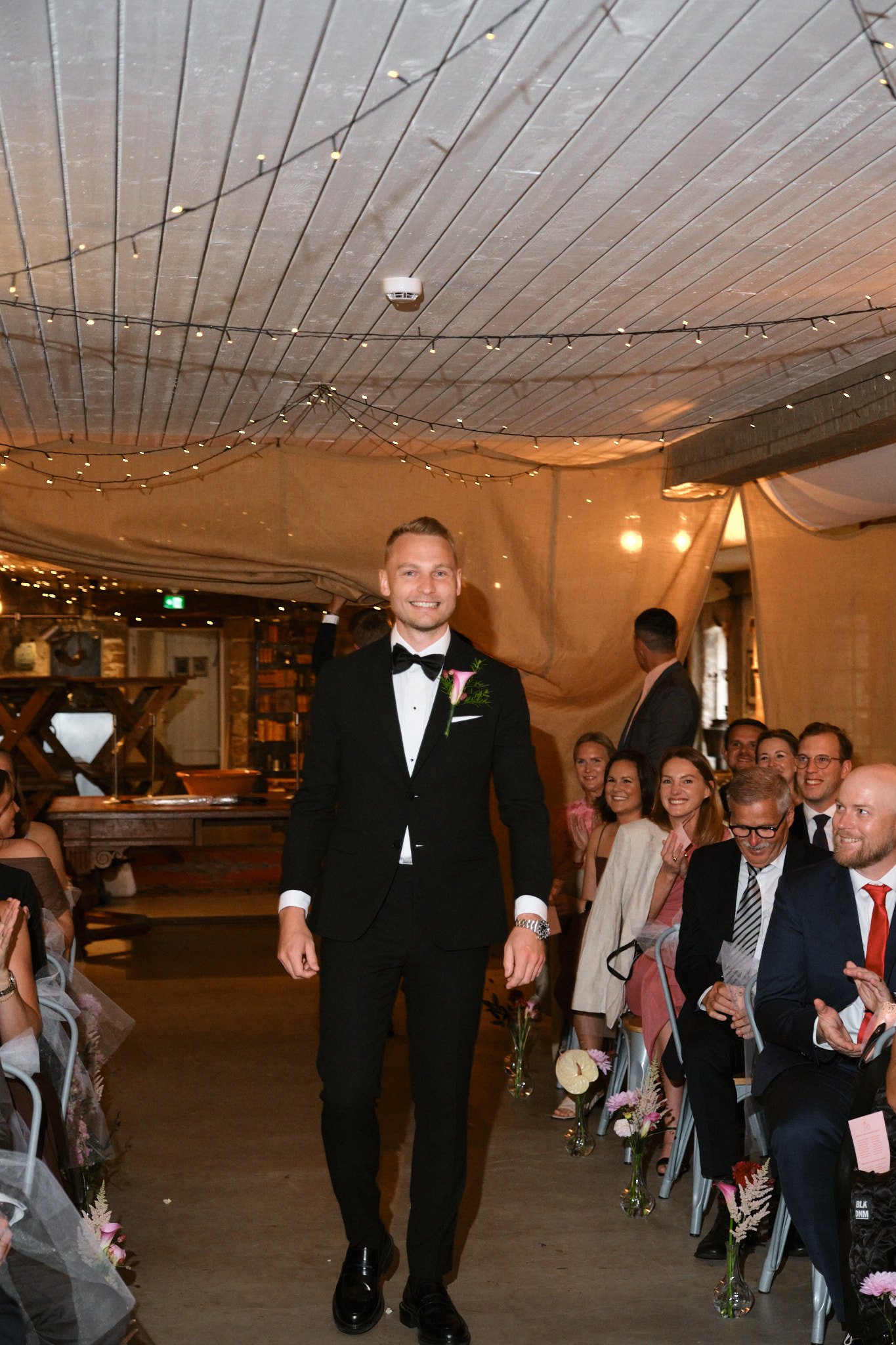 Man i svart tuxedo med blommor på bröstet, går nerför gången vid ett bröllop, omgiven av gäster som sitter och ler.
