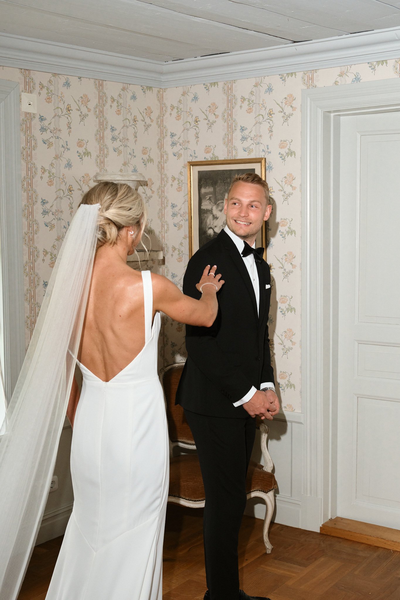 En brud i vit klänning med rygg utsatt, och en brudgubbe i svart tuxedo, står i ett rum med blommig tapet, har ett glädjefyllt uttryck.