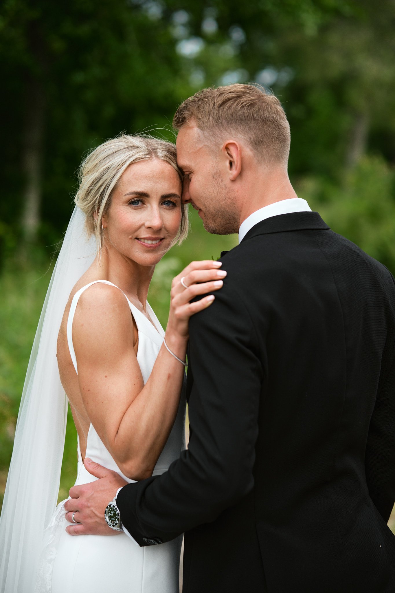 En nygift brud och brudgum står nära varandra utomhus, med brudens hand vilande på brudgummens axel, båda i formella kläder, i en grön park.