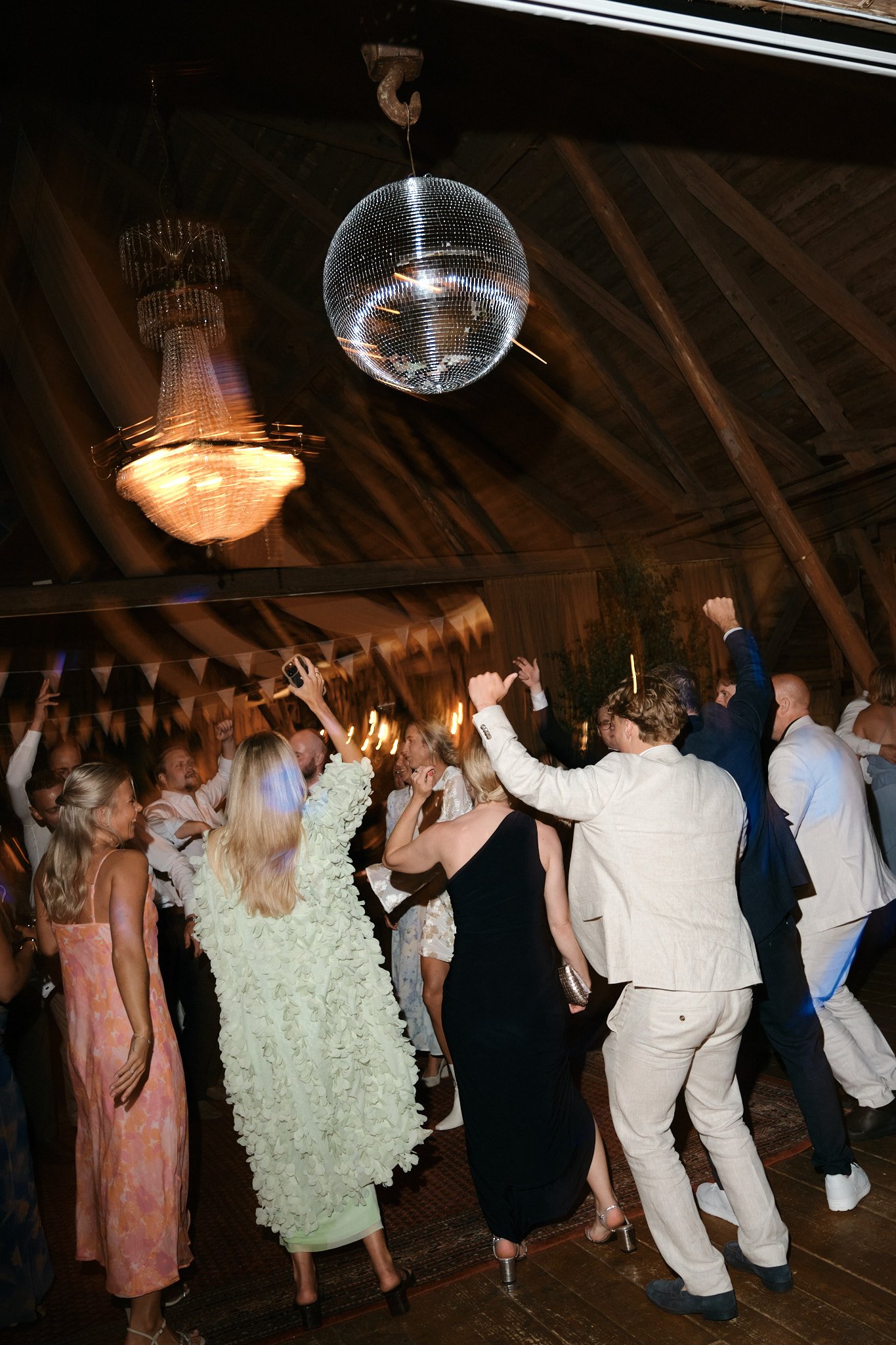 Dansande människor vid fest i en träbyggnad med disco-ball och kristallkrona.
