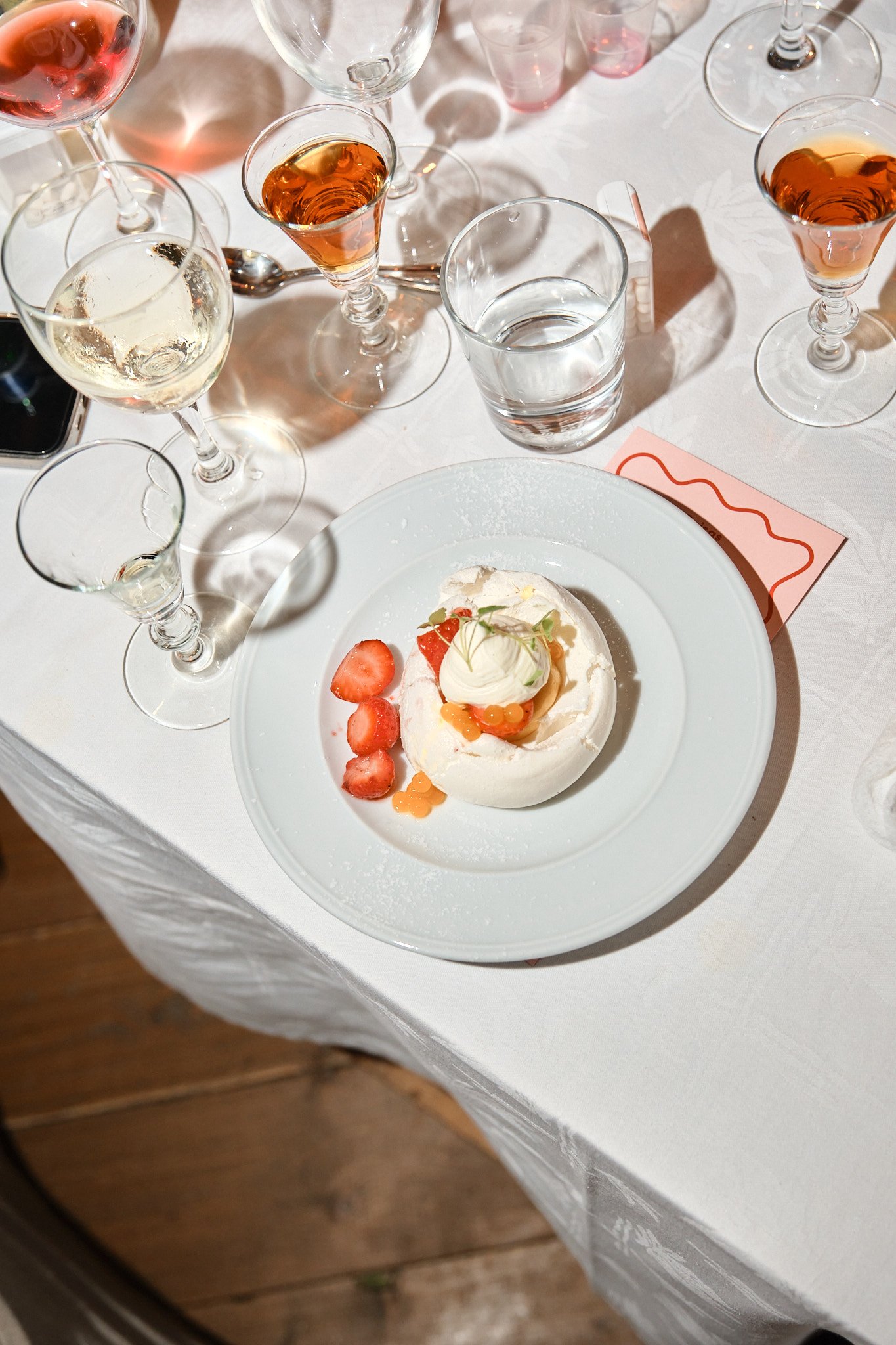 En vit tallrik med en efterrätt som innehåller jordgubbar, en liten rund grädde- eller glassboll, och dekorativa element, omgiven av olika glas med drycker, inklusive vin och mousserande vin, på ett vitt dukat bord.